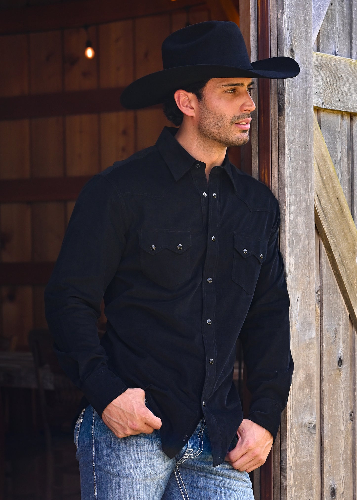 Man standing in a doorway wearing a black cowboy hat and a black snap shirt. 