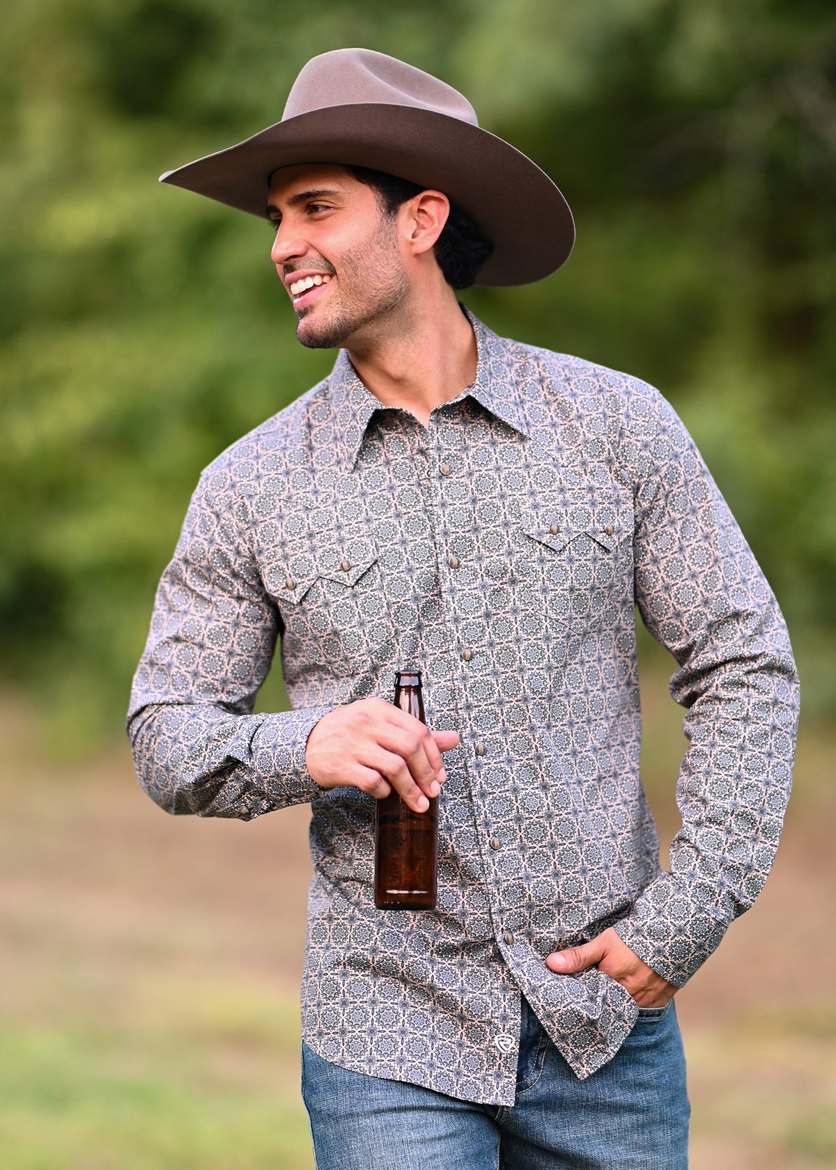 A man wearing a brown cowboy hat and a gray button-down shirt with a white geometric pattern, holding a beer bottle against an outdoor background.