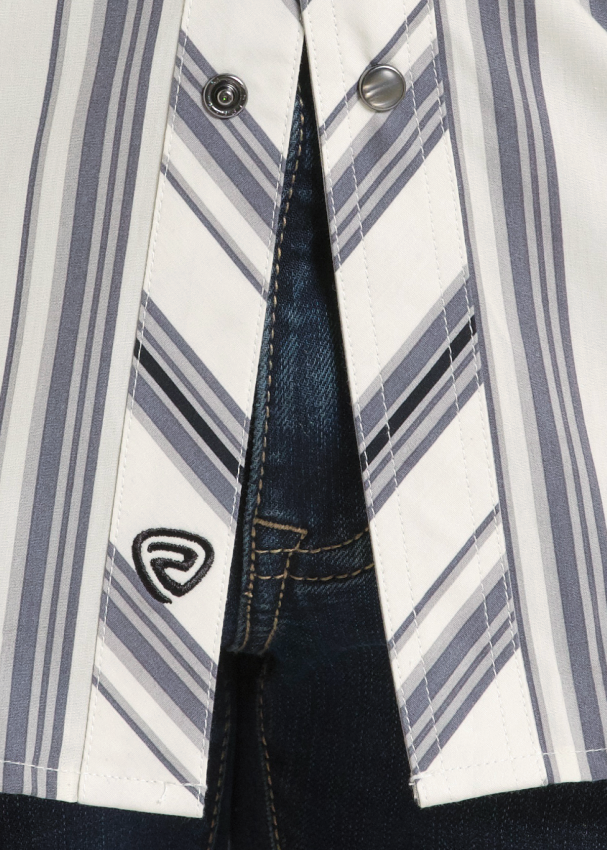 Close-up of a striped button-up shirt with white, gray, and navy vertical and diagonal stripes, worn open over dark blue denim jeans. A small black embroidered shield logo is visible on the shirt pocket area.