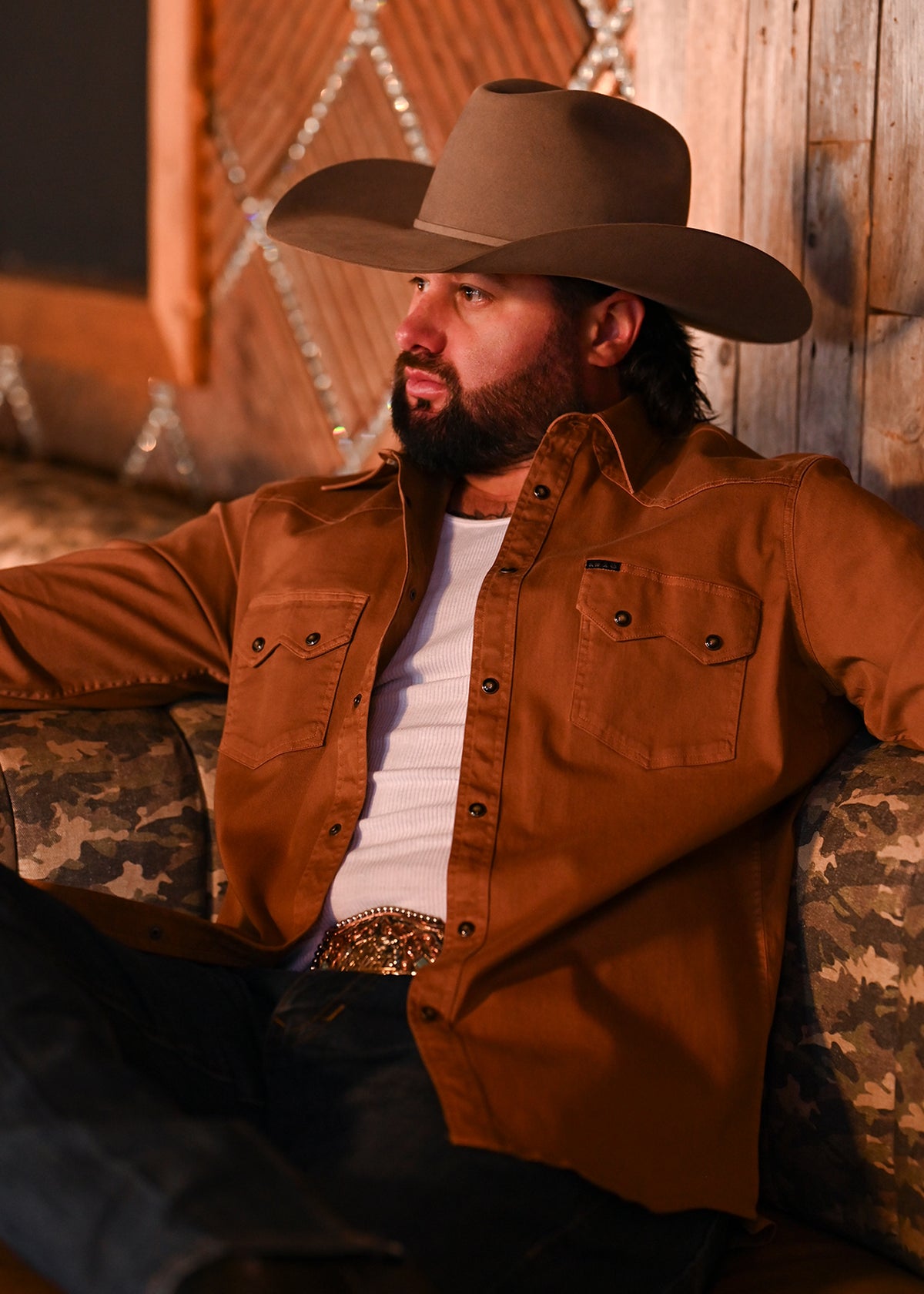 Koe Wetzel wearing a cowboy hat, a button-up shirt over a white t-shirt, and jeans, sitting on a couch in an indoor setting.