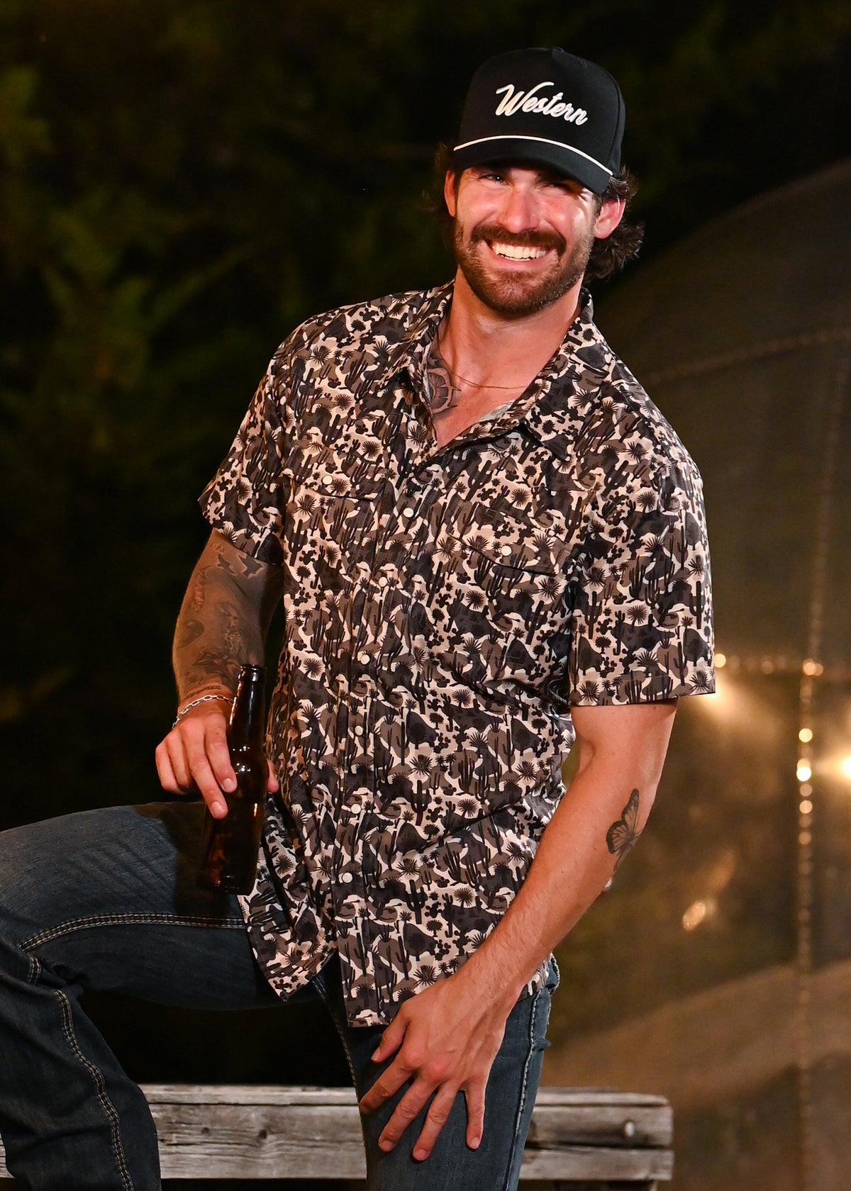 A smiling man wearing a black baseball cap, a black-and-beige camo patterned short-sleeve snap shirt, and dark jeans, posed outdoors at night.