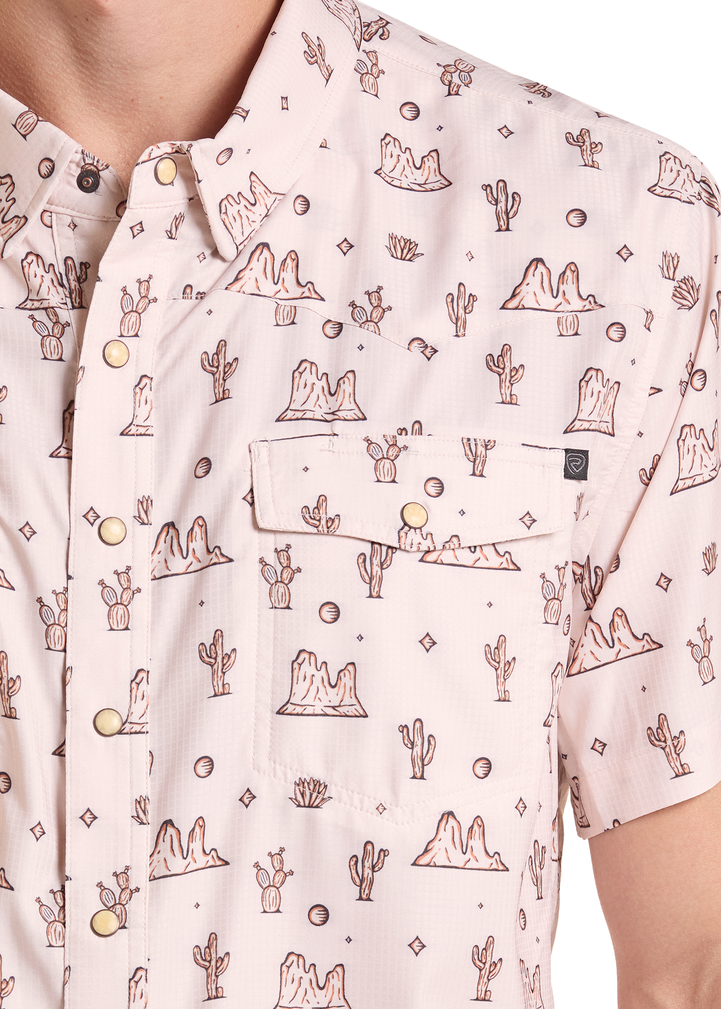 A close-up of a short-sleeve button-up shirt featuring a light pink base with a brown desert cactus and rock pattern, accented by gold-tone snap buttons and a chest pocket.