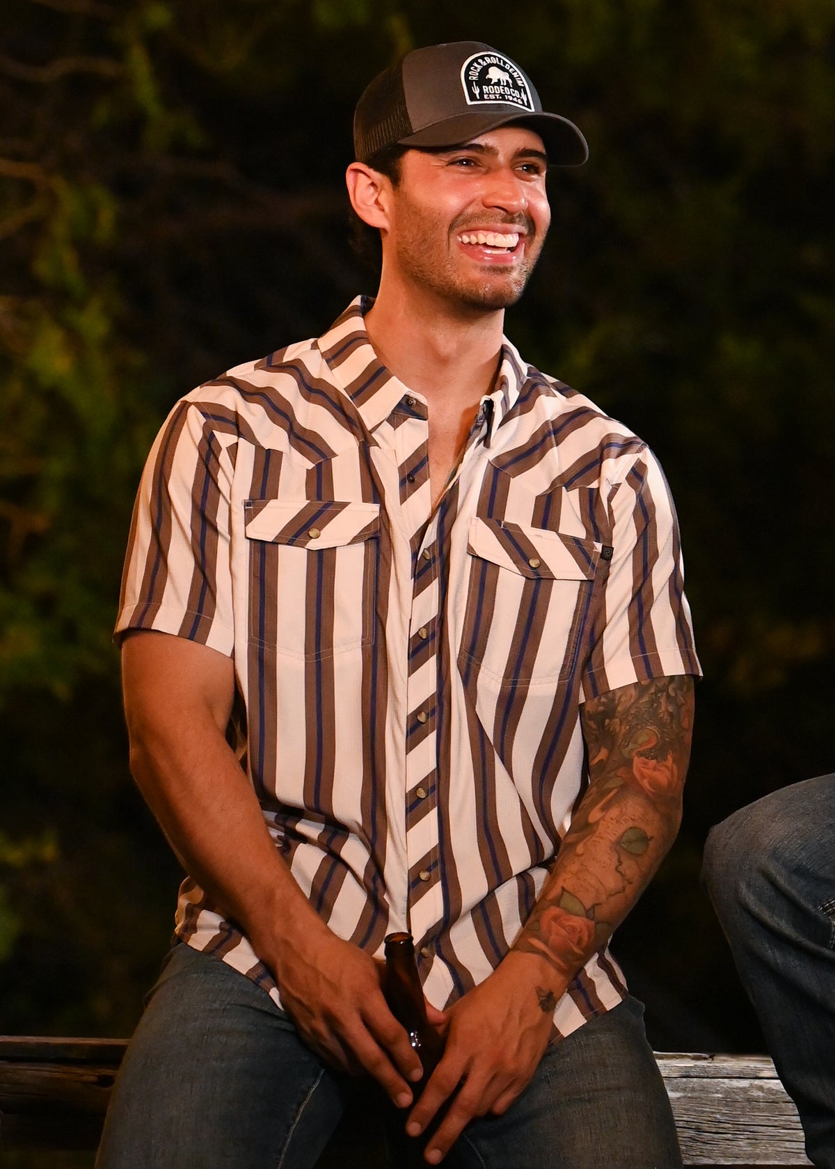 A man wearing a brown and cream striped short-sleeve button-up shirt, blue jeans, and a dark baseball cap with a circular logo, sitting outdoors at night.
