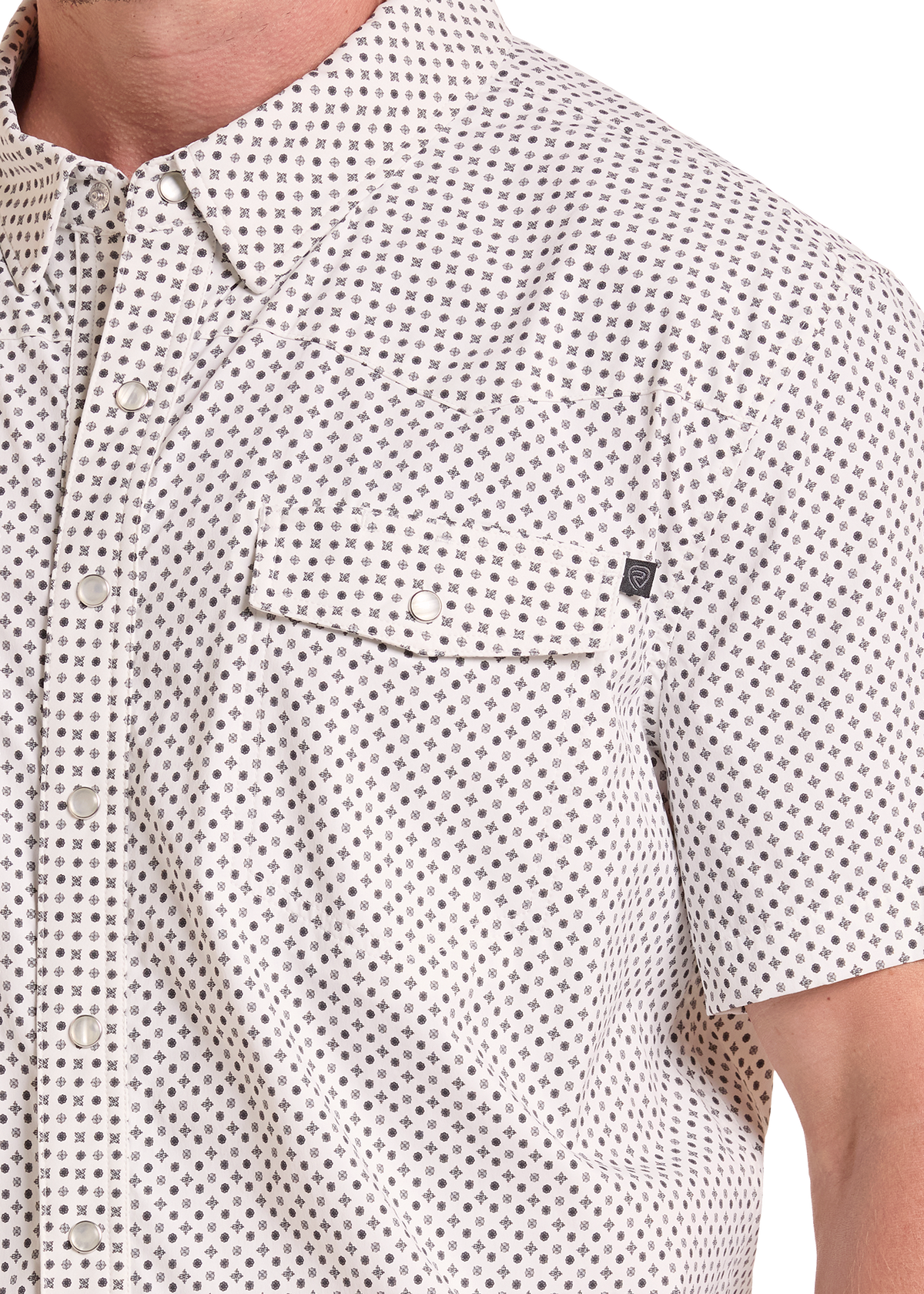 Close-up of a man wearing a short-sleeve button-up shirt with a small repeating geometric pattern in dark gray on an off-white base. The shirt features a classic collar, a chest pocket with a snap tab, and a small black brand tag on the pocket.