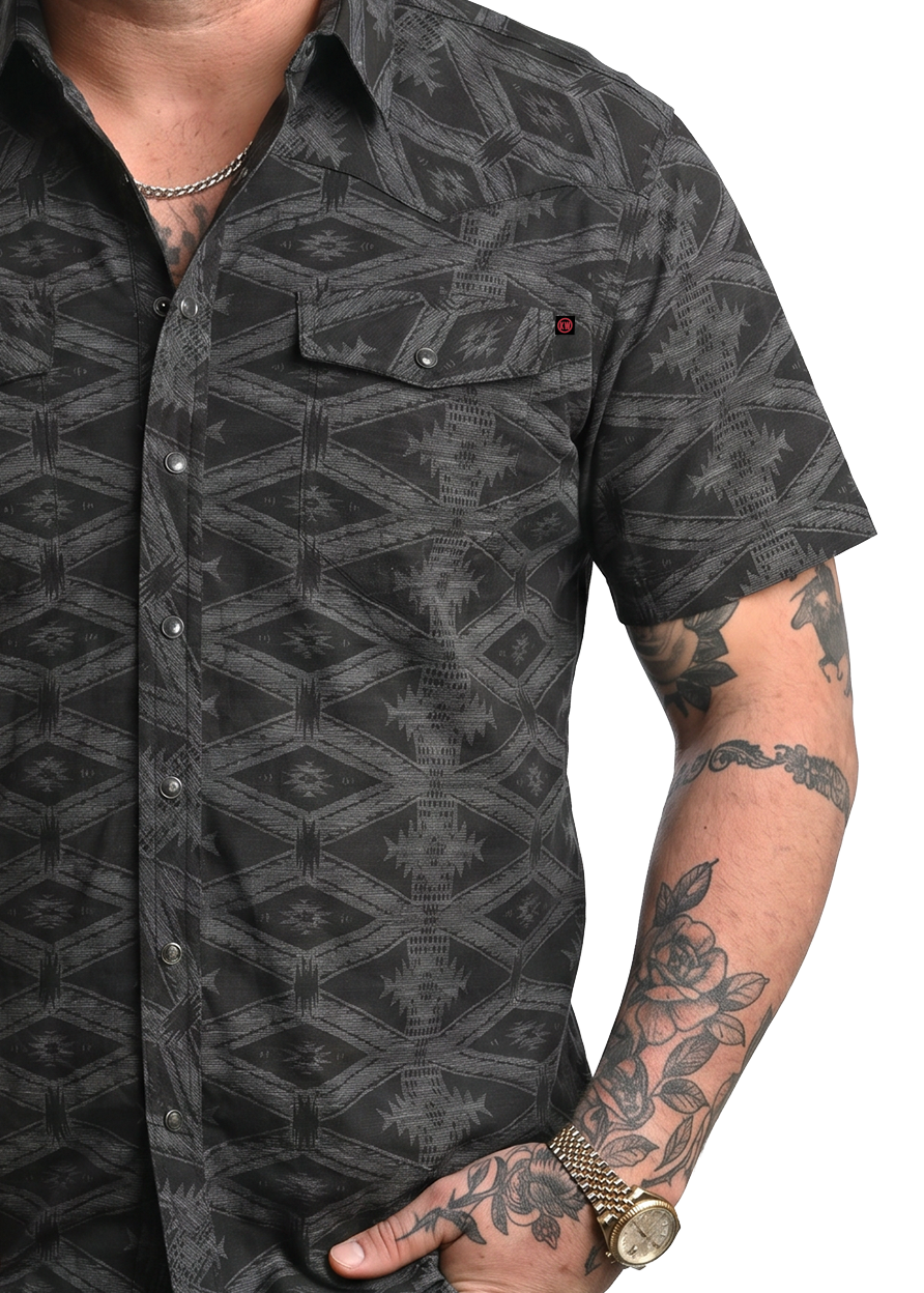 Man wearing a black patterned shirt with tattoos on arms, on a white background