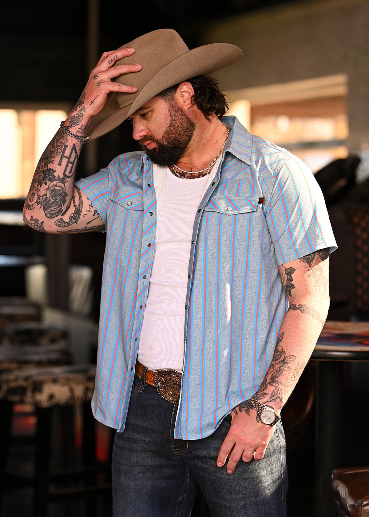 Koe Wetzel wearing a grey and blue stripe shirt unbuttoned with jeans a a cowboy hat. 