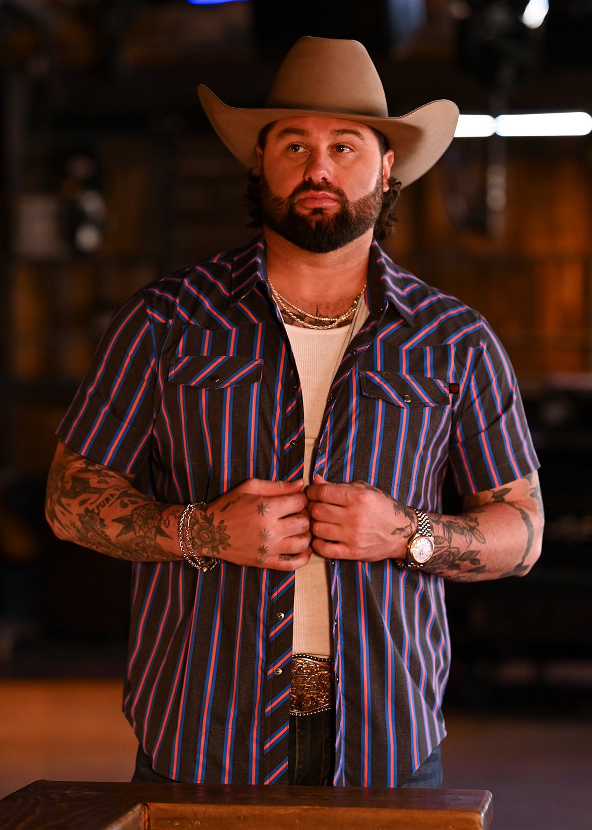 Koe Wetzel wearing a cowboy hat, a vertically striped short-sleeve snap shirt over a white undershirt standing in a bar.