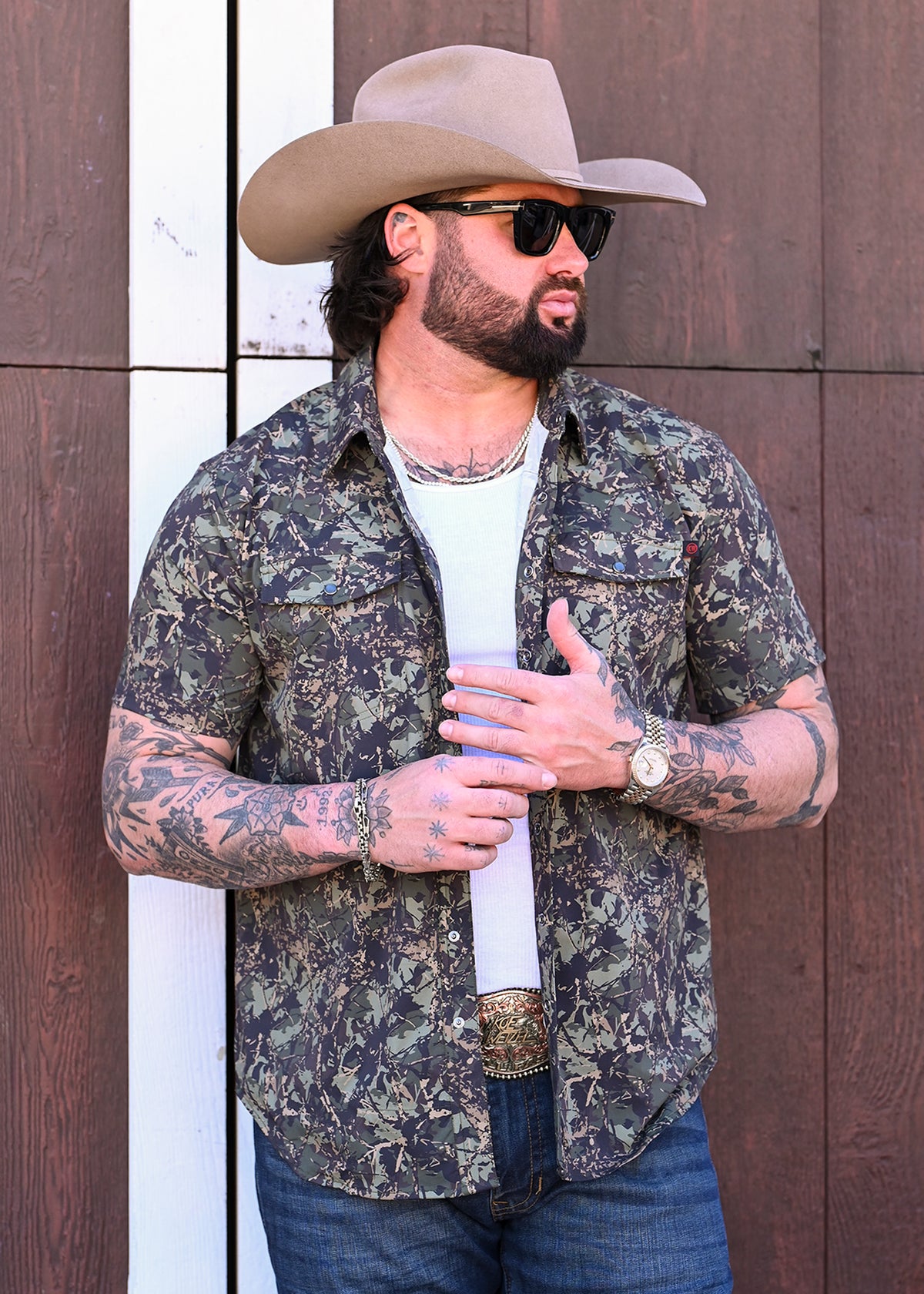Koe Wetzel wearing a camo performance shirt unbuttoned over a white shirt with jeans and a cowboy hat. 