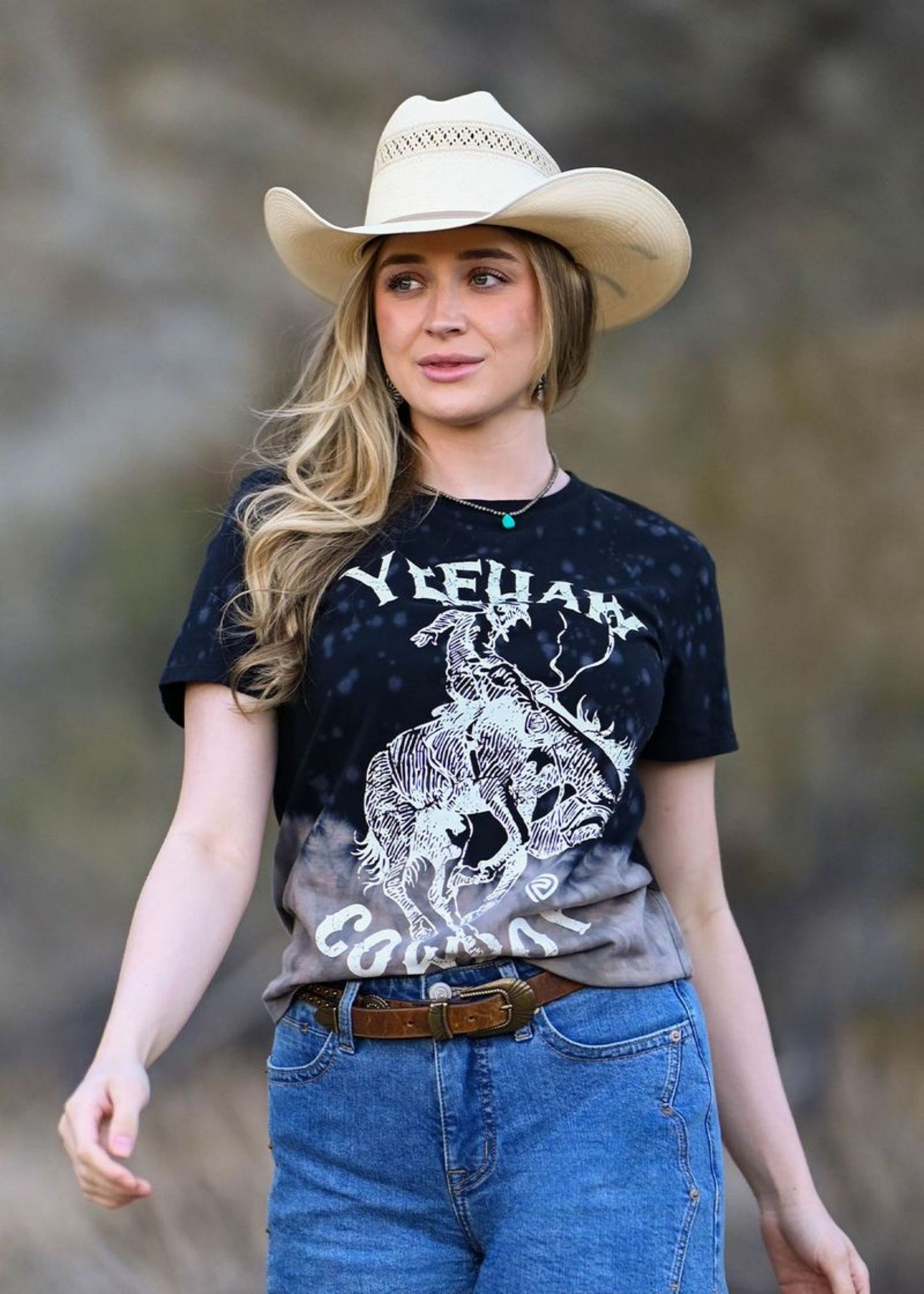 Woman wearing a black t-shirt with graphic design and a cowboy hat outdoors.