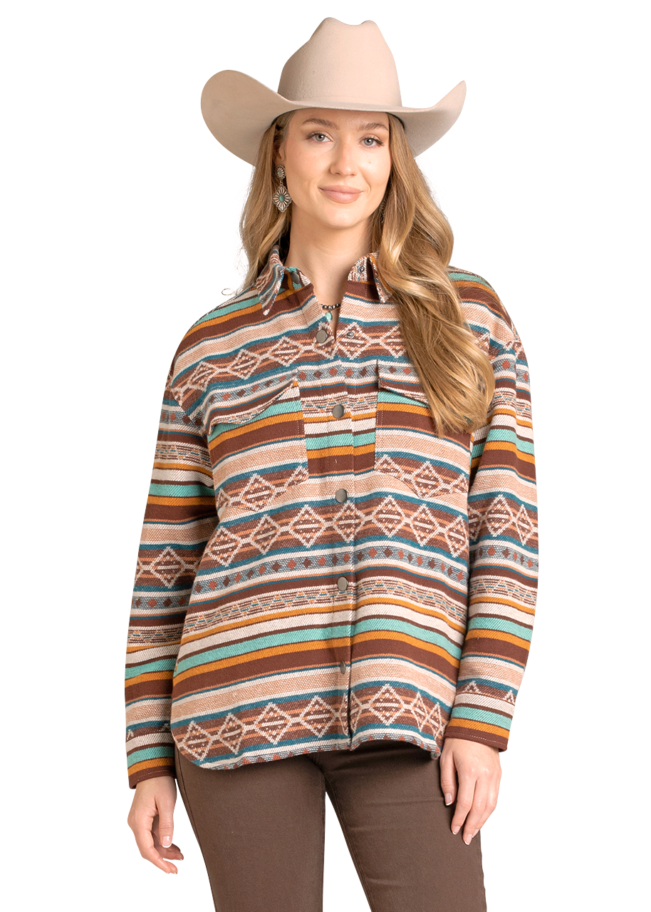 A woman smiles against a plain background, wearing the Rock & Roll Denim Multicolored Aztec Shacket with earthy geometric designs, a beige cowboy hat, and brown pants.