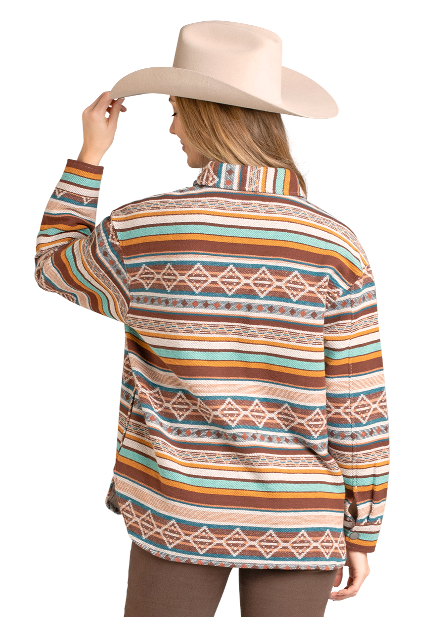 A woman in a beige cowboy hat and brown pants stands facing away, touching her hat. She wears the Rock & Roll Denim Multicolored Aztec Shacket, adding a casual Southwestern layer to her look.