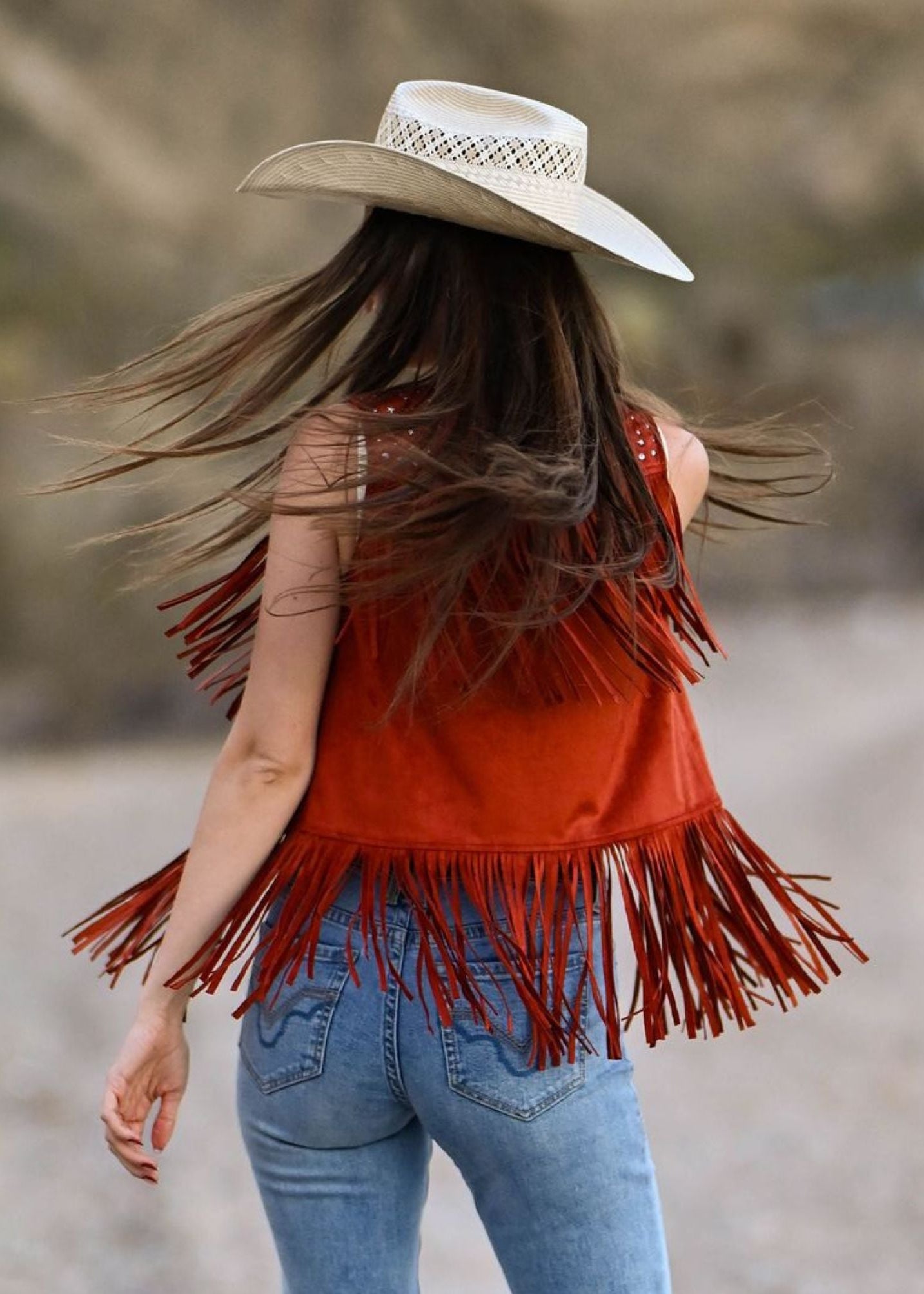 Person wearing a red fringe vest and white cowboy hat with long hair blowing in the wind.
