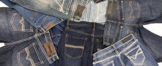 A close-up flat lay of multiple denim jeans spread out, showing different washes from light blue to dark blue with contrasting white stitching and brown leather patch pockets.