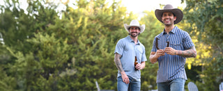Two men wearing Western shirts outdoors. The man in the foreground wears a cowboy hat, a blue-and-white polo, and jeans holding a horseshoe.