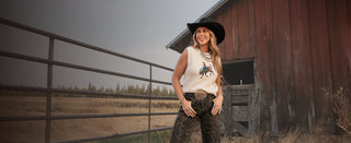 West Desperado standing in a field next to a barn wearing a sleeveless sweater and leopard jeans. 