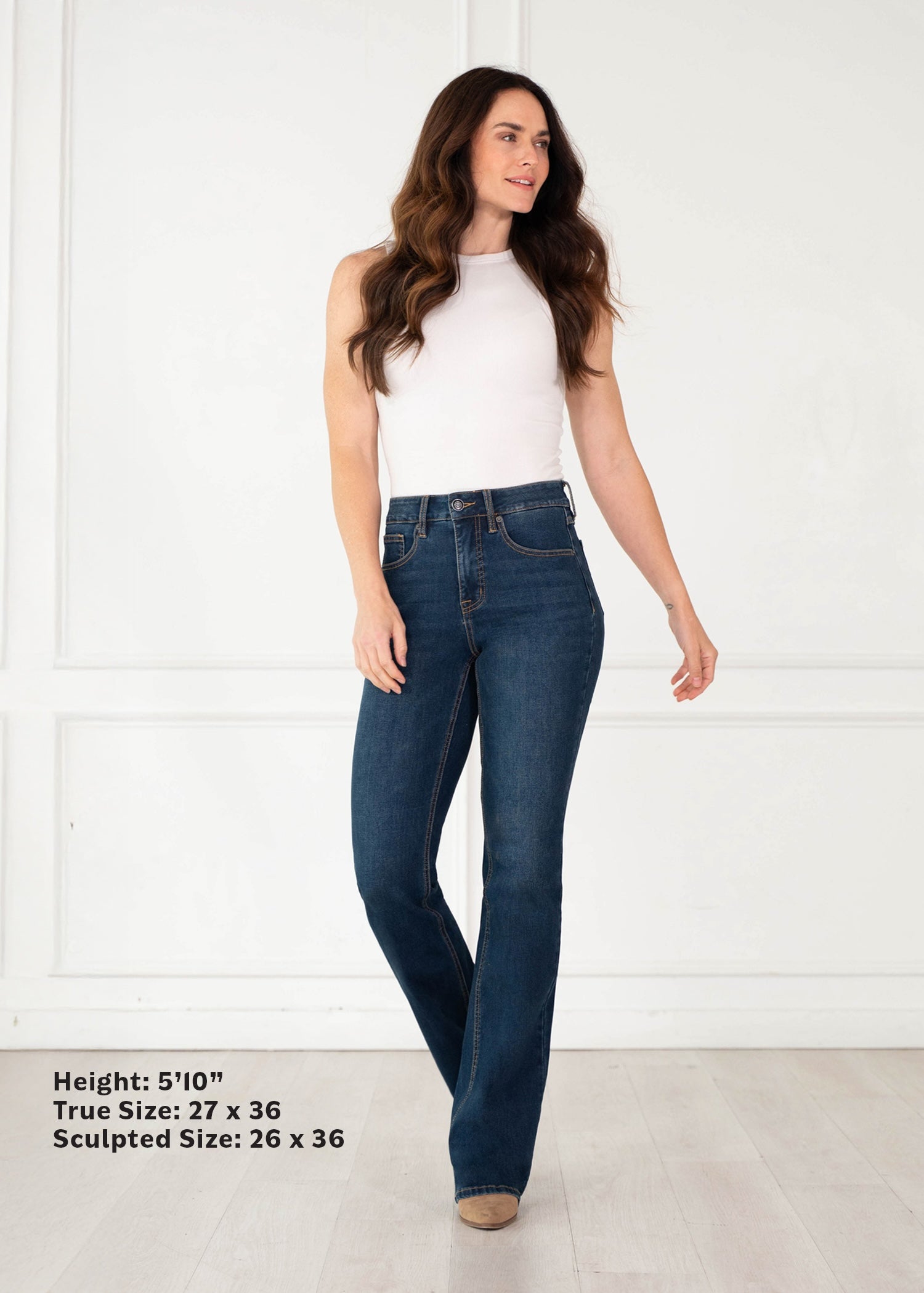 A woman modeling dark blue high-waisted flare jeans with a white sleeveless top, standing against a white paneled wall. Text in the bottom-left corner lists height and true/sculpted sizes.