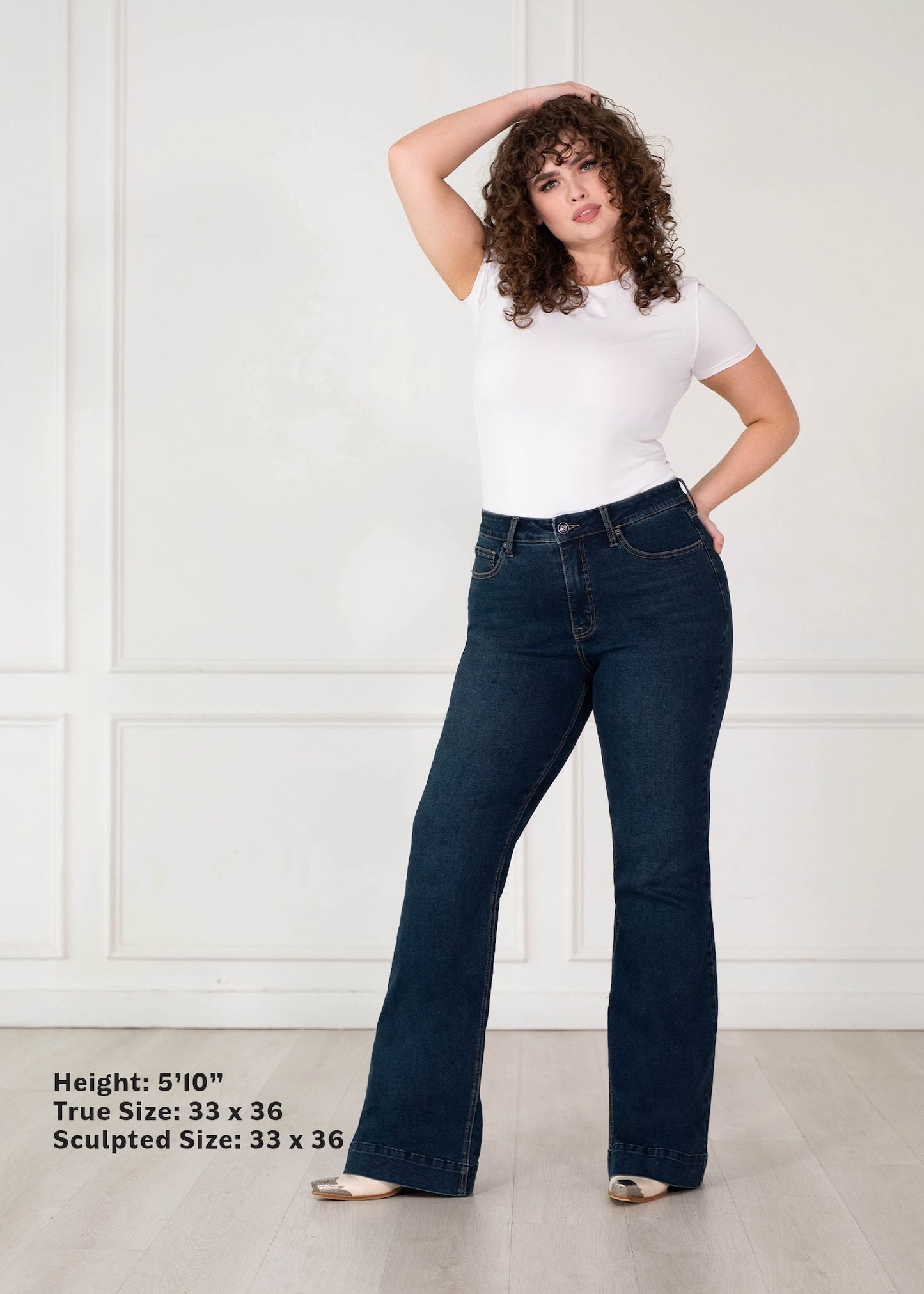 A woman modeling dark blue flare jeans with a white t-shirt, standing in a white paneled room. Text in the bottom-left corner lists height and true/sculpted sizes.