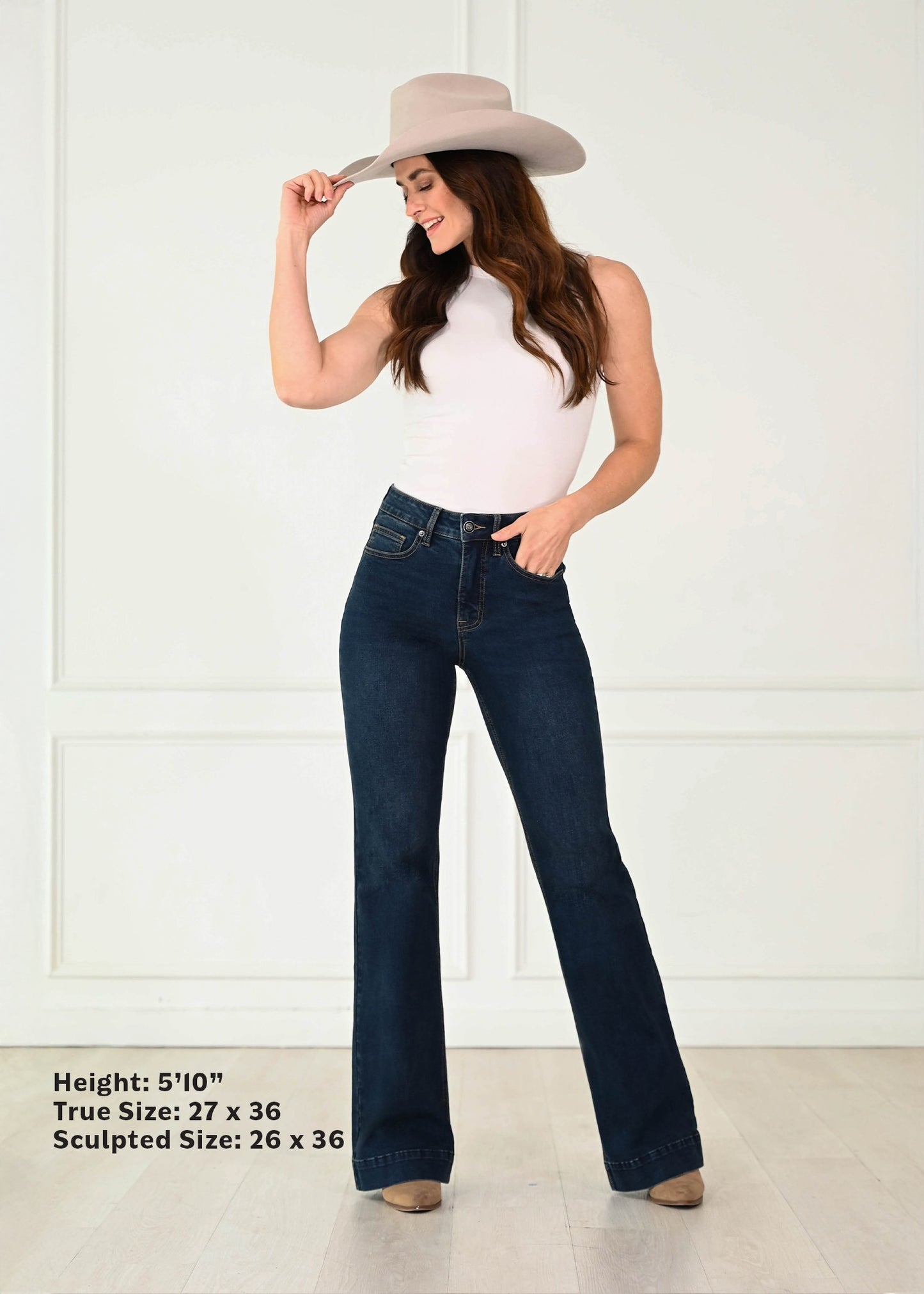 A woman modeling dark blue flare jeans with a white sleeveless top and a beige cowboy hat, standing against a white paneled wall. Text in the bottom left corner lists height and sizing information.