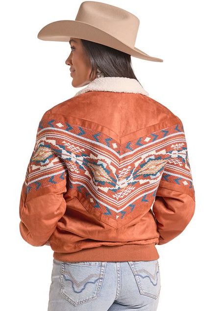 A woman shown from the back wearing a rust-orange western-style bomber jacket with a colorful geometric pattern across the shoulders and upper back, a cream sherpa collar.