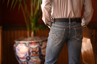 Person wearing a beige shirt and blue jeans standing indoors with a decorative vase and plant in the background.