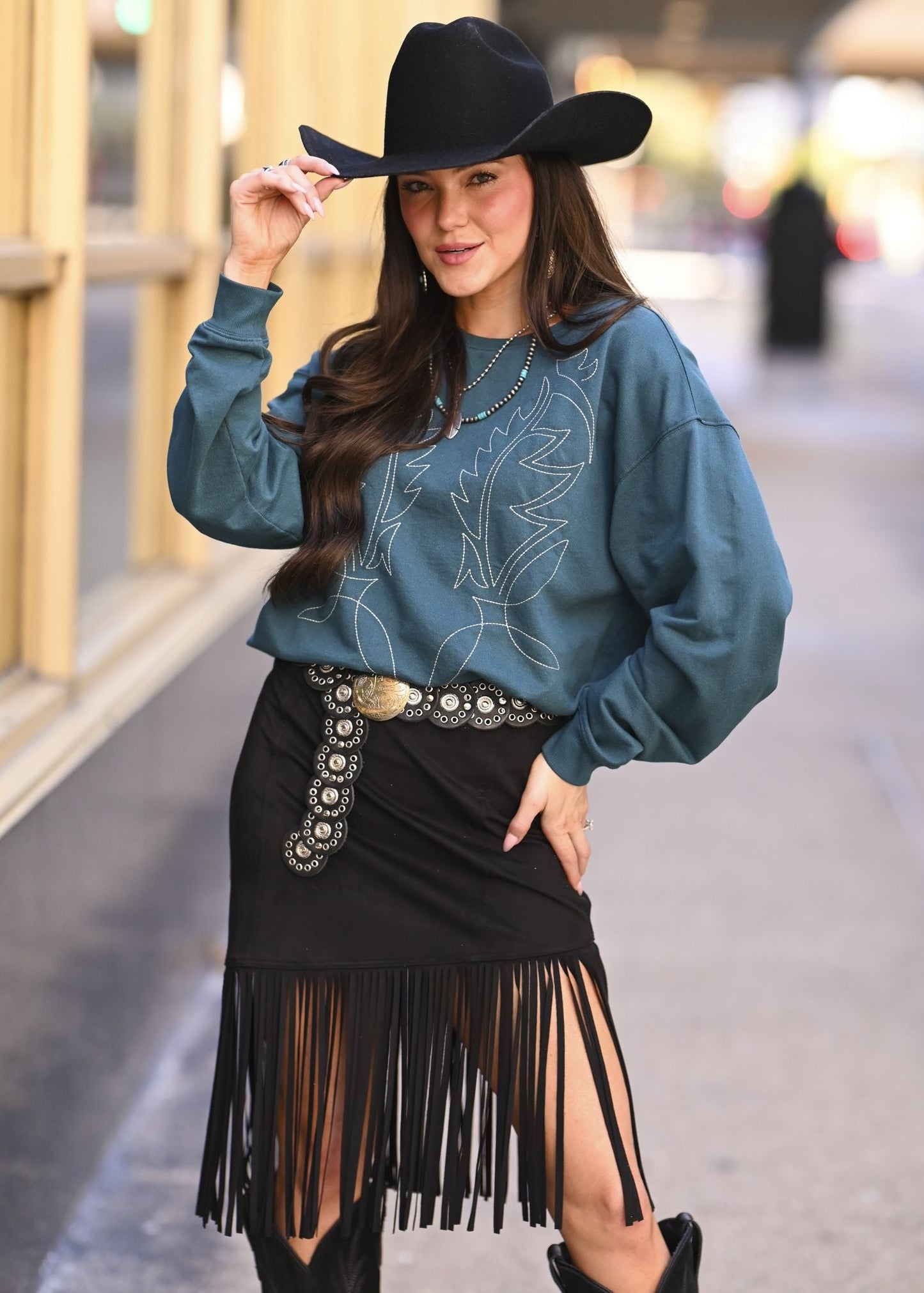 Woman wearing a teal sweater and black fringe skirt with a cowboy hat outdoors.