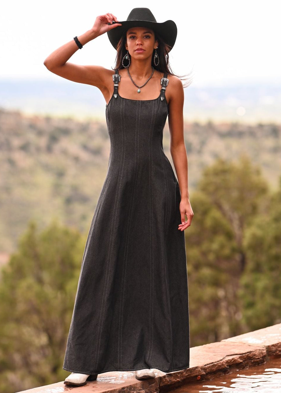 Woman in a black dress and hat standing on a ledge with a scenic background
