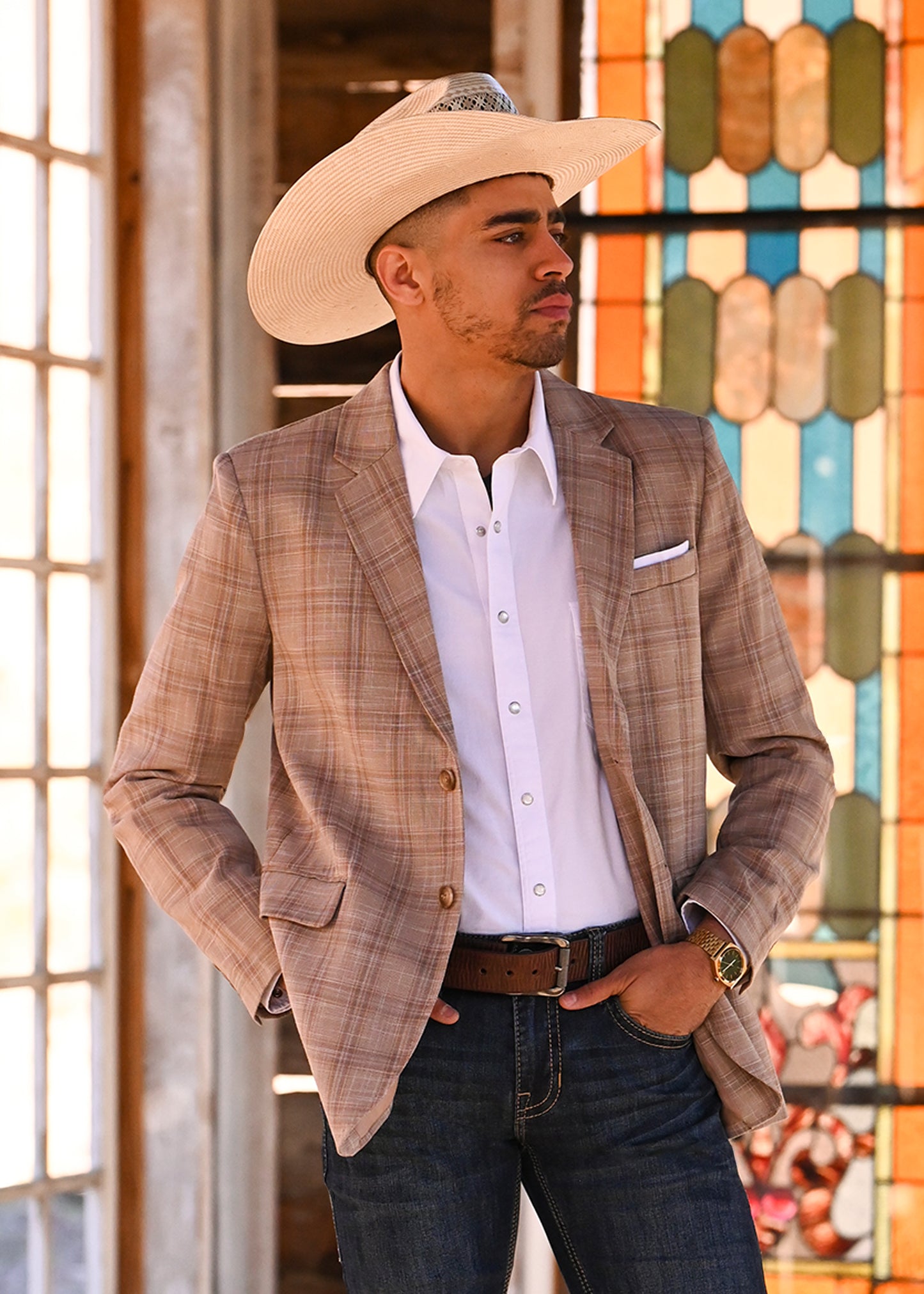 Man wearing a plaid blazer, white shirt, and cowboy hat in front of a colorful stained glass window.