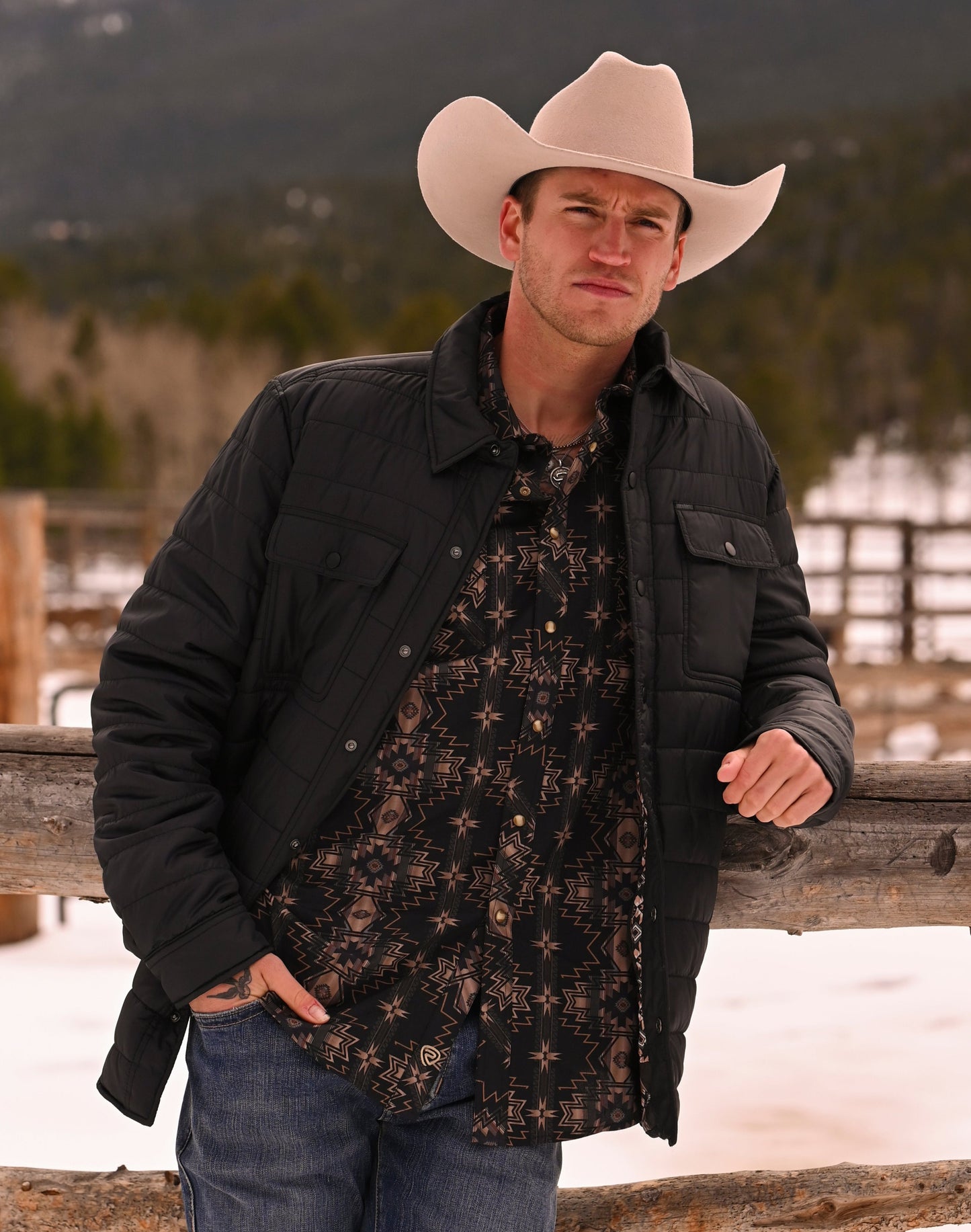 A man in a light cowboy hat and Rock & Roll Denim Puffer Print Lined Shirt Jacket leans against a wooden fence outdoors, surrounded by snow, mountains, and trees.