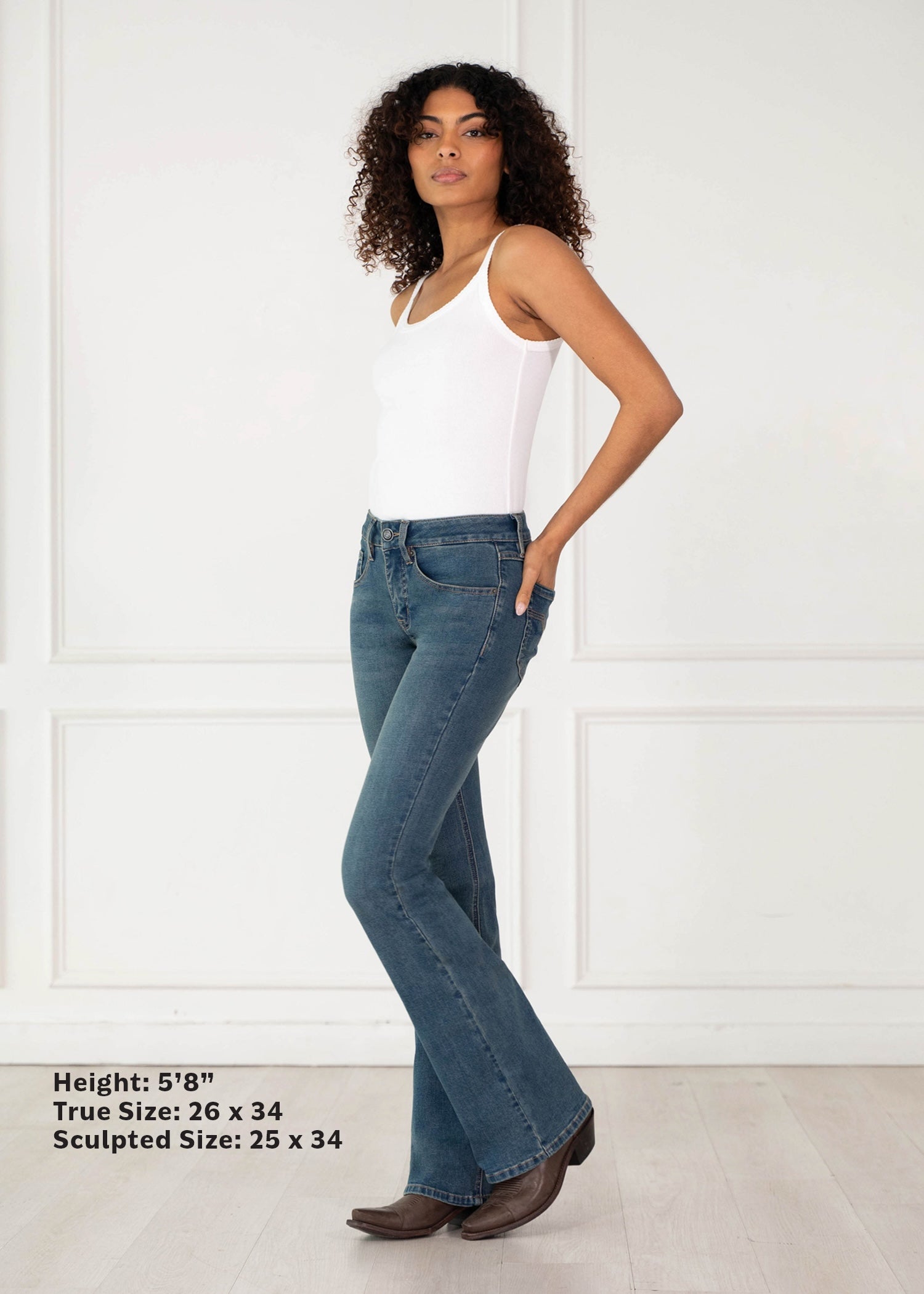 A woman modeling blue flare jeans with a white tank top and brown boots, standing in a bright white room with paneling. Text in the lower left lists height, true size, and sculpted size.