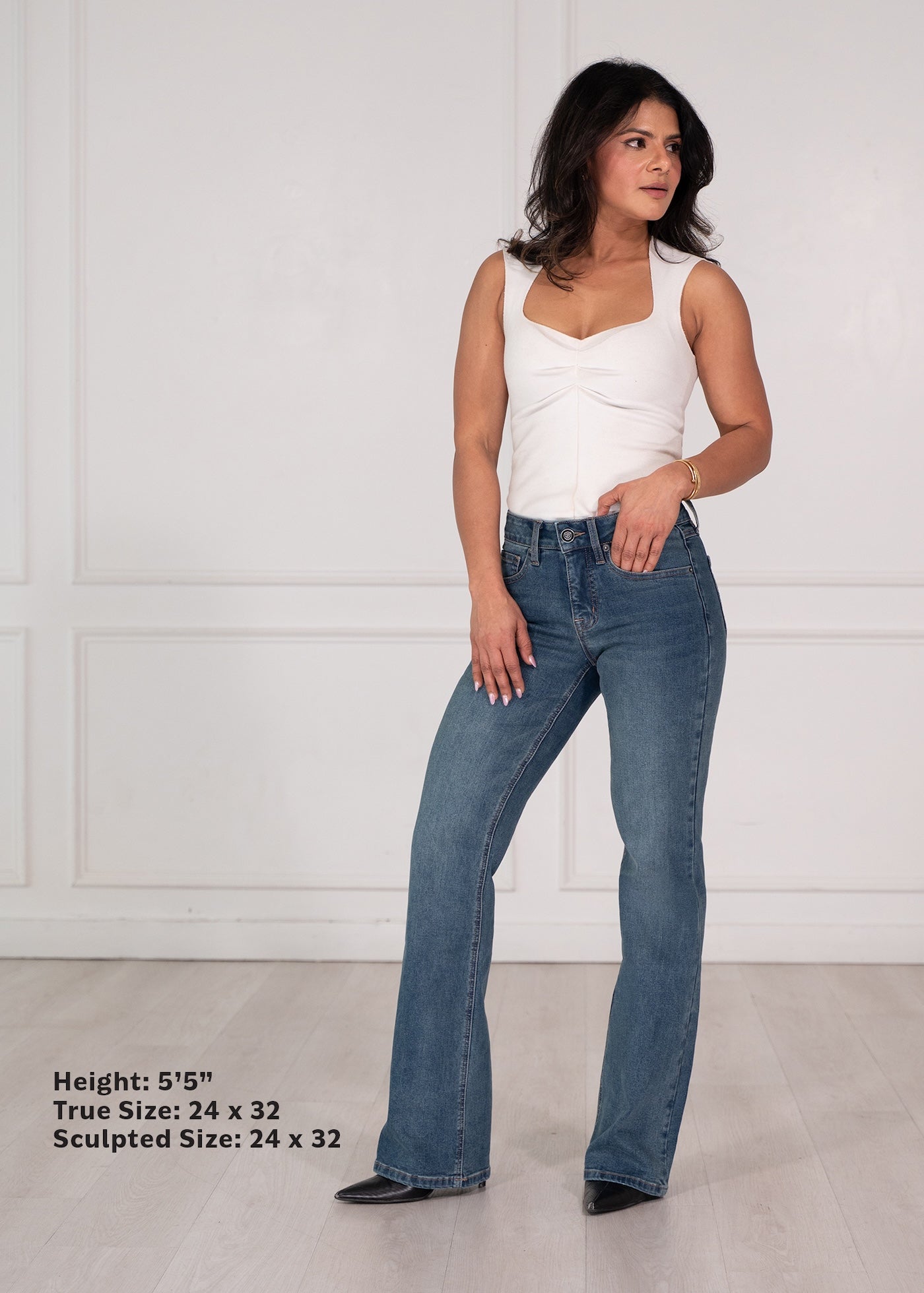 A woman modeling a pair of medium-wash blue flare jeans with a white sleeveless top, standing against a white paneled wall. Text in the bottom-left corner lists height and true/sculpted sizes.