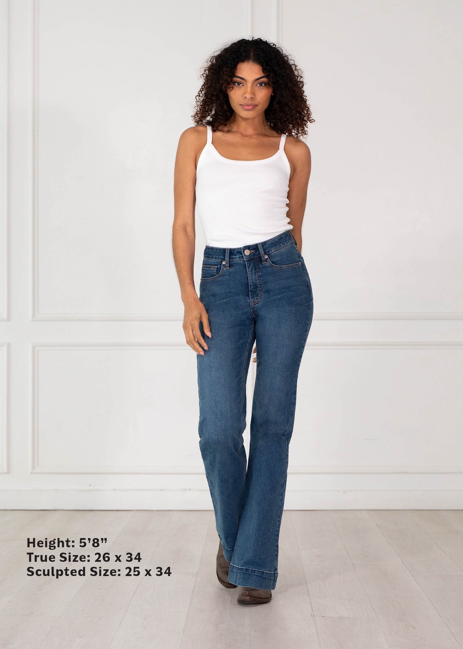 A model wearing blue high-waisted flare jeans and a white tank top stands against a white paneled wall. Black text in the bottom-left corner lists height and sizing information for the jeans.