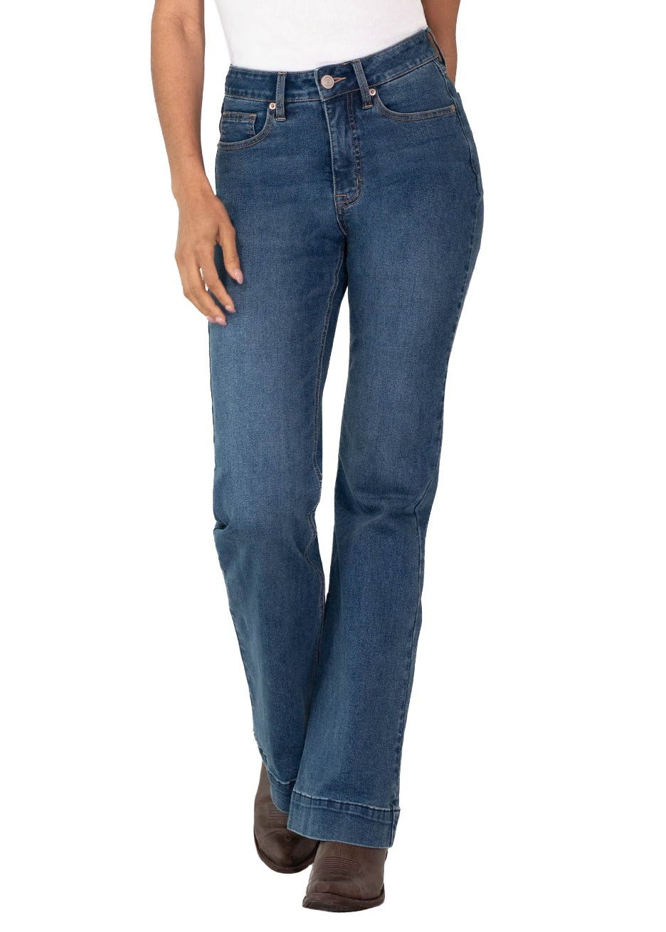 A woman modeling a pair of medium-wash blue jeans with a white tank top and brown boots, standing against a plain white background.