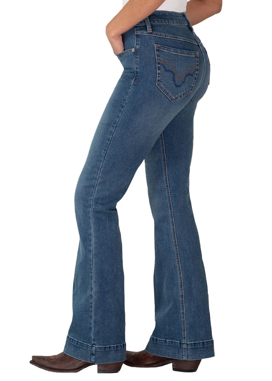 A woman modeling a pair of blue flare jeans, styled with a white tank top, a beige cowboy hat, and brown cowboy boots, shown from a side-back profile against a white background.