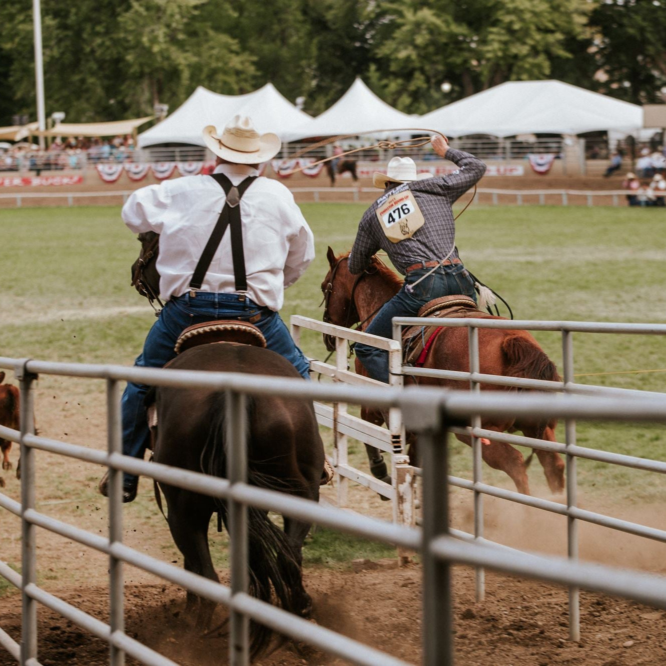 Two cowboys on horseback in a rodeo arena with spectators and tents in the background.