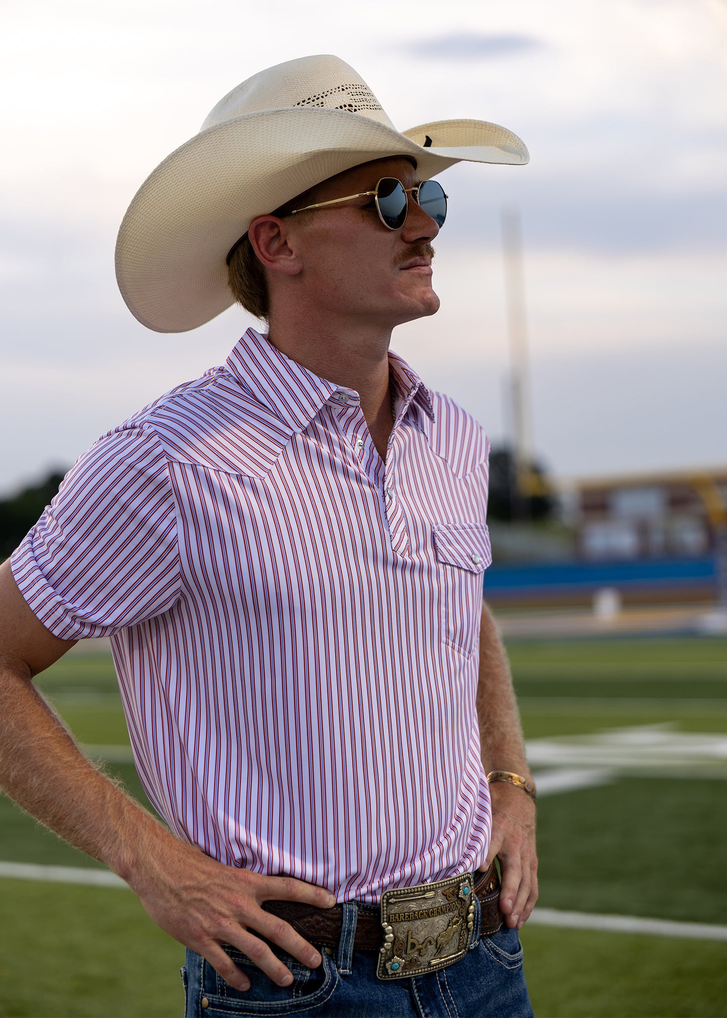 A man wearing a white cowboy hat and a red and white vertically striped short-sleeve button-up shirt, standing on a sports field.