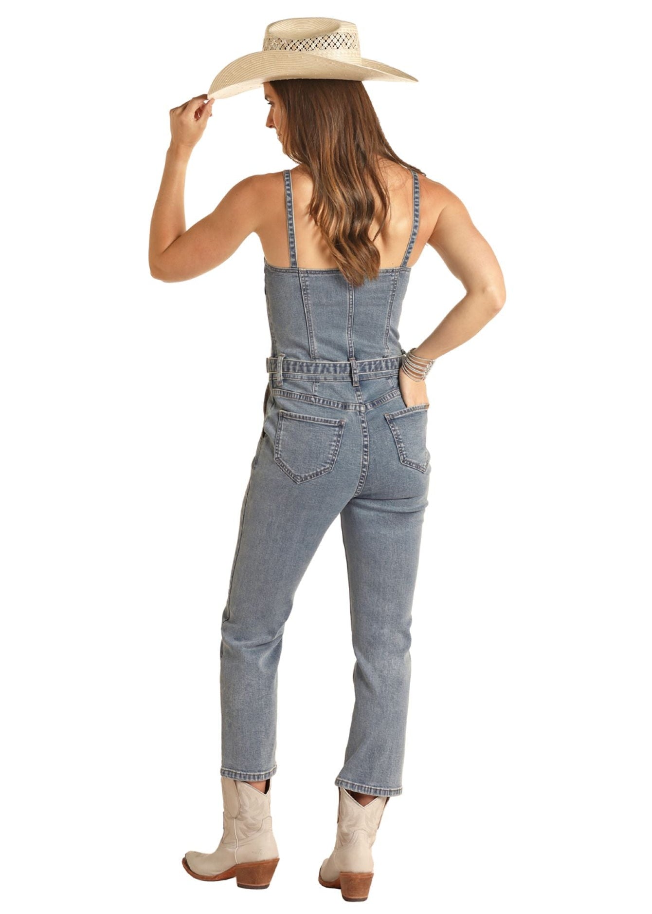 A woman faces away wearing the Rock & Roll Denim Front Seams Denim Jumpsuit, paired with a white cowboy hat and light boots. She holds her hat brim with one hand and rests the other on her hip.