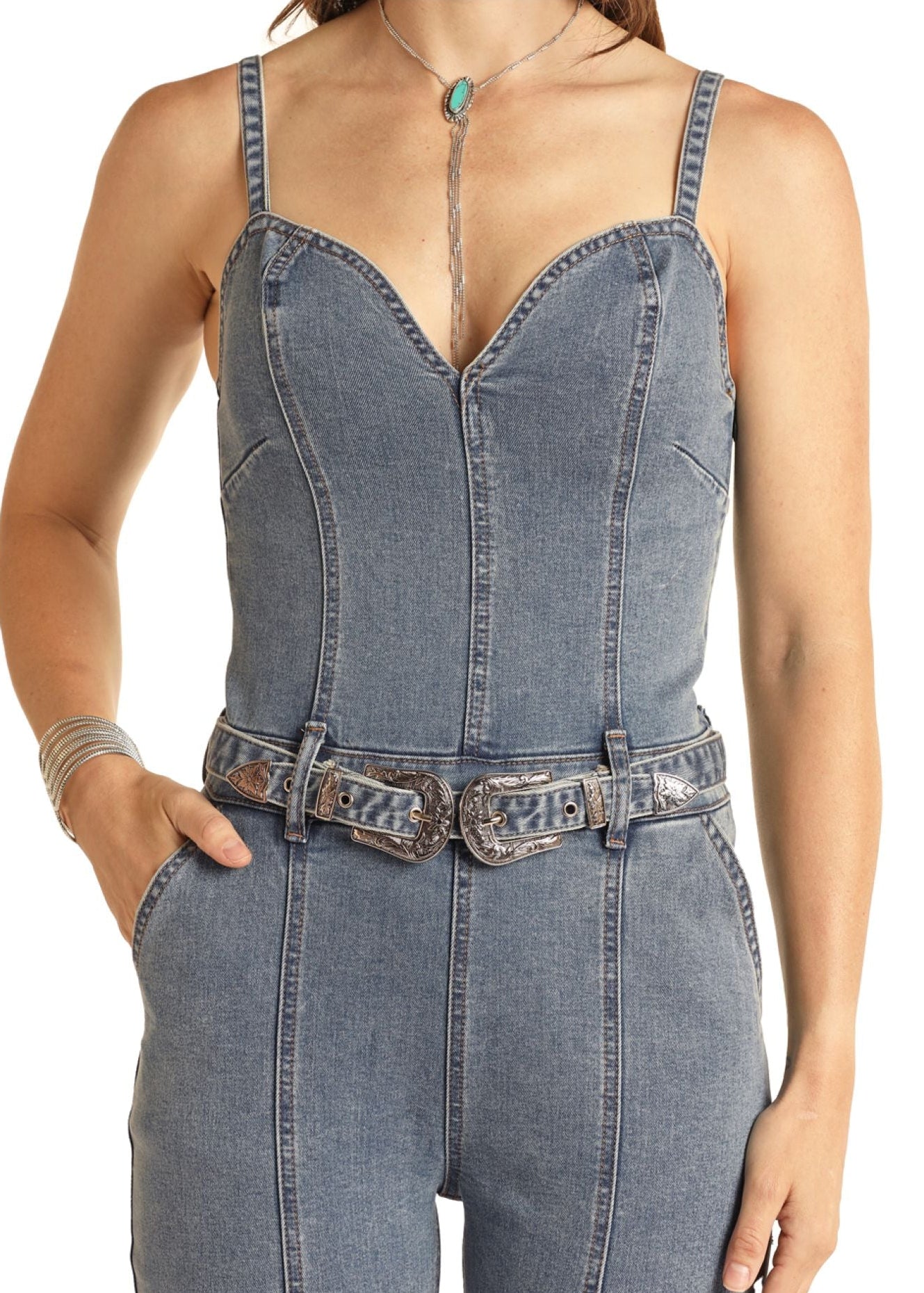 A person wears the Rock & Roll Denim Front Seams Denim Jumpsuit with a flattering fit and sweetheart neckline, styled with a silver double belt, turquoise pendant necklace, one hand in their pocket, and a silver bracelet.