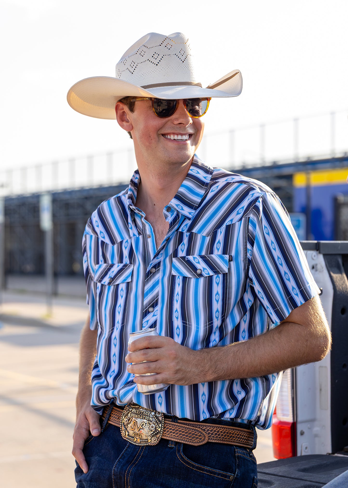 A smiling man wearing a white cowboy hat, sunglasses, a blue and white striped short-sleeve button-up shirt, and a brown belt with a large buckle, holding a drink outdoors near a truck.