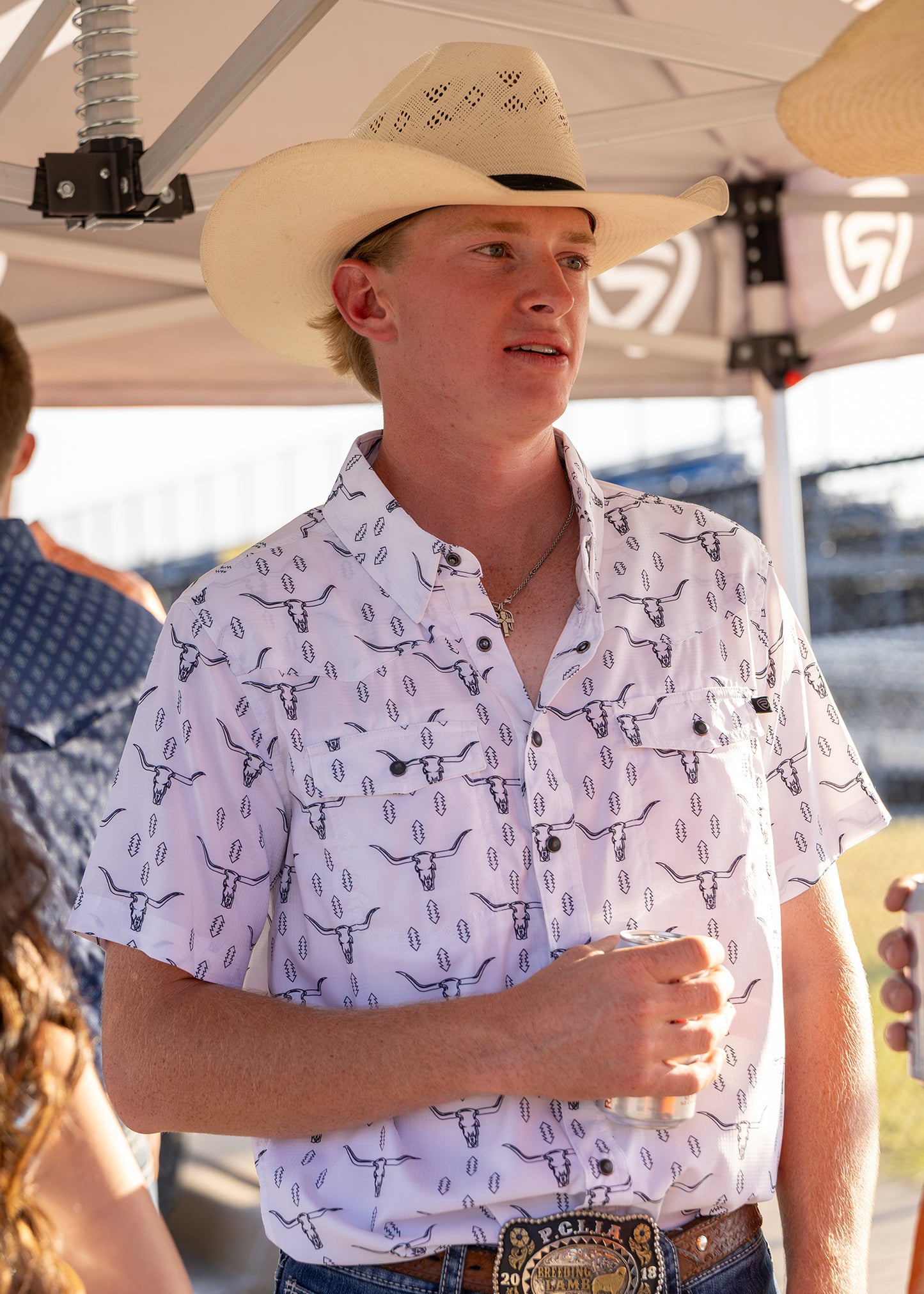 Man wearing a white button down shirt with steer skulls on it, a white cowboy hat and blue jeans standing under a canopy with a drink in his hand. 