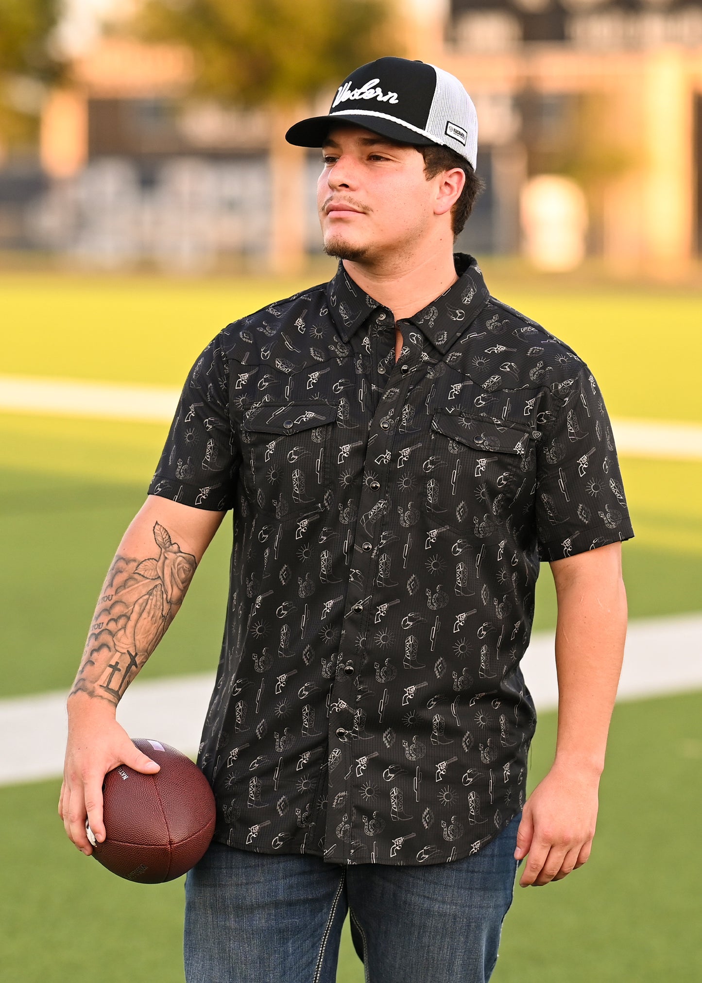 A man in a Rock & Roll Denim TEK Western Desert Print Short Sleeve Shirt, jeans, and a black-and-white cap stands on a football field holding a football. Blurred green trees and buildings appear in the background.