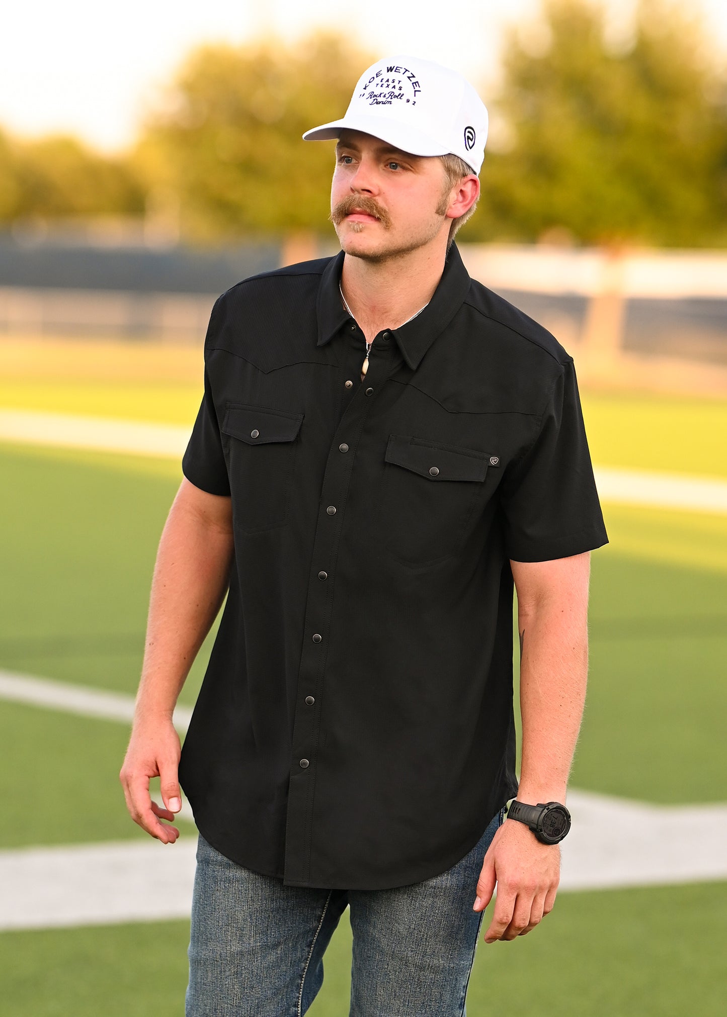 A man with a mustache, wearing a white cap, jeans, black watch, and the Rock & Roll Denim TEK Western Black Short Sleeve Shirt stands on a sports field with green grass and blurred trees in the background.