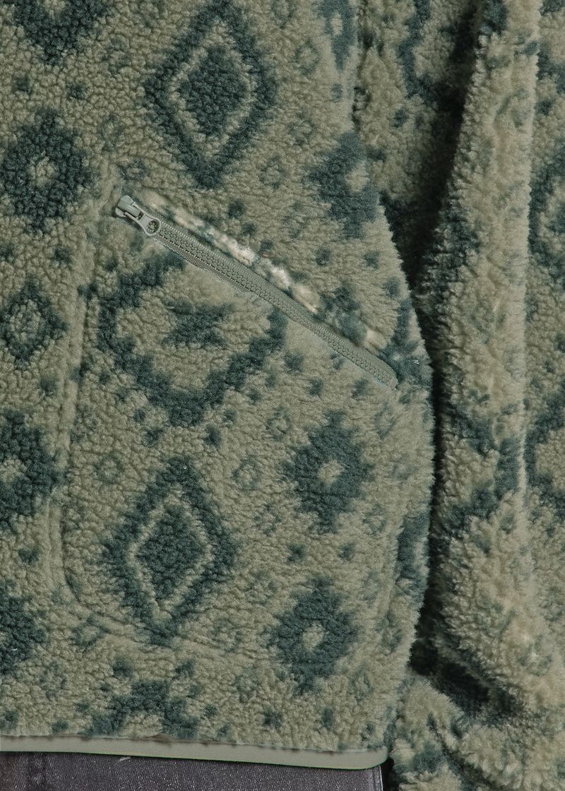 Close-up of a sherpa fleece jacket with a geometric diamond pattern in sage green and dark gray, featuring a diagonal zippered pocket.