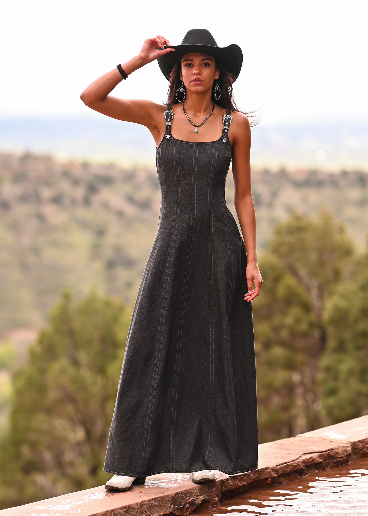 A woman wears the Rock & Roll Denim Buckle Strap Denim Maxi Dress and a black wide-brimmed hat while standing outdoors on a stone ledge, holding her hat with one hand. Trees and hills in the background add a Western-inspired flair.