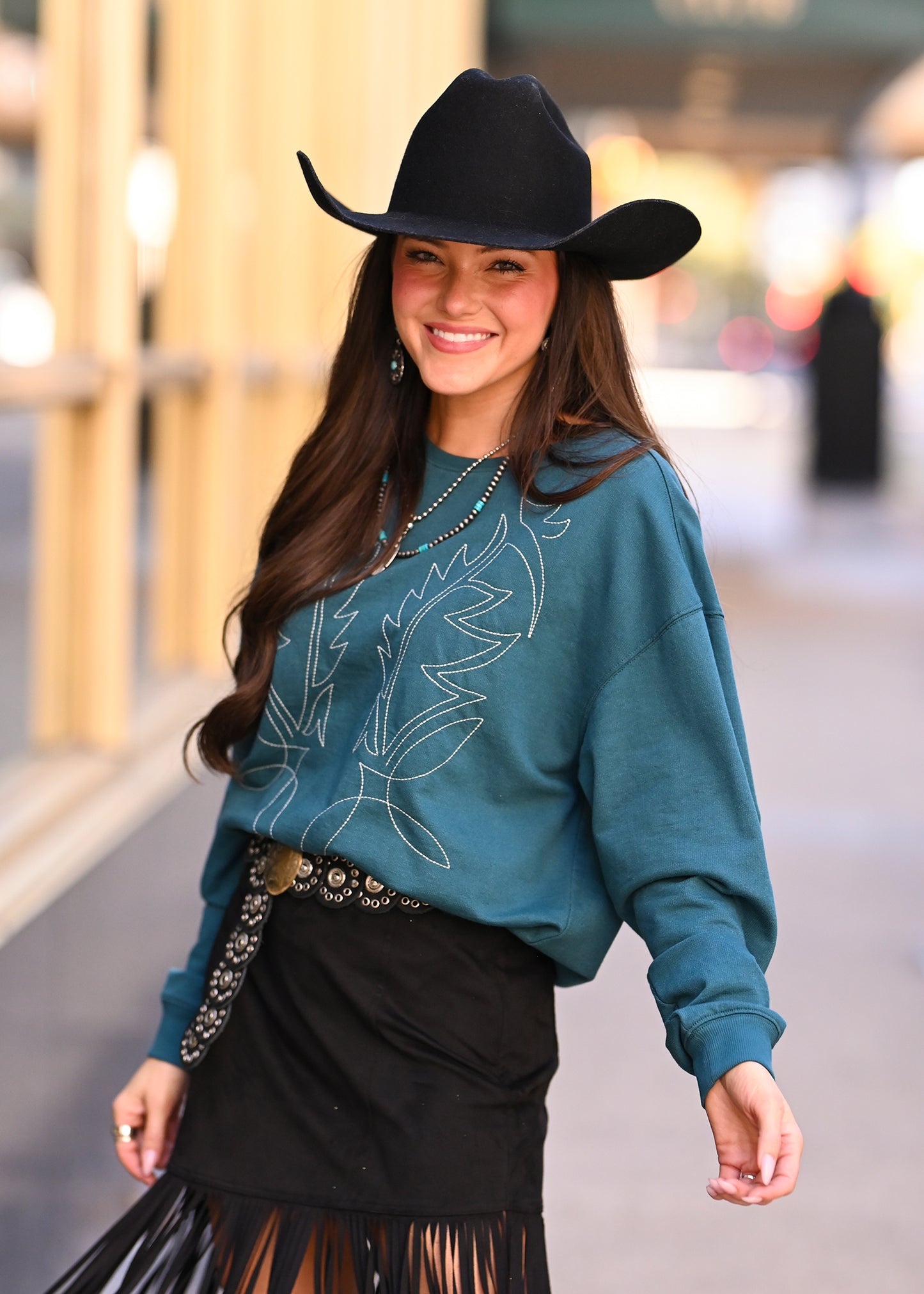 A smiling young woman stands on a city sidewalk wearing a black cowboy hat, black fringed skirt, decorated belt, and the Boot Stitch Pullover Sweatshirt by Rock & Roll Denim. Blurred buildings and lights appear in the background.