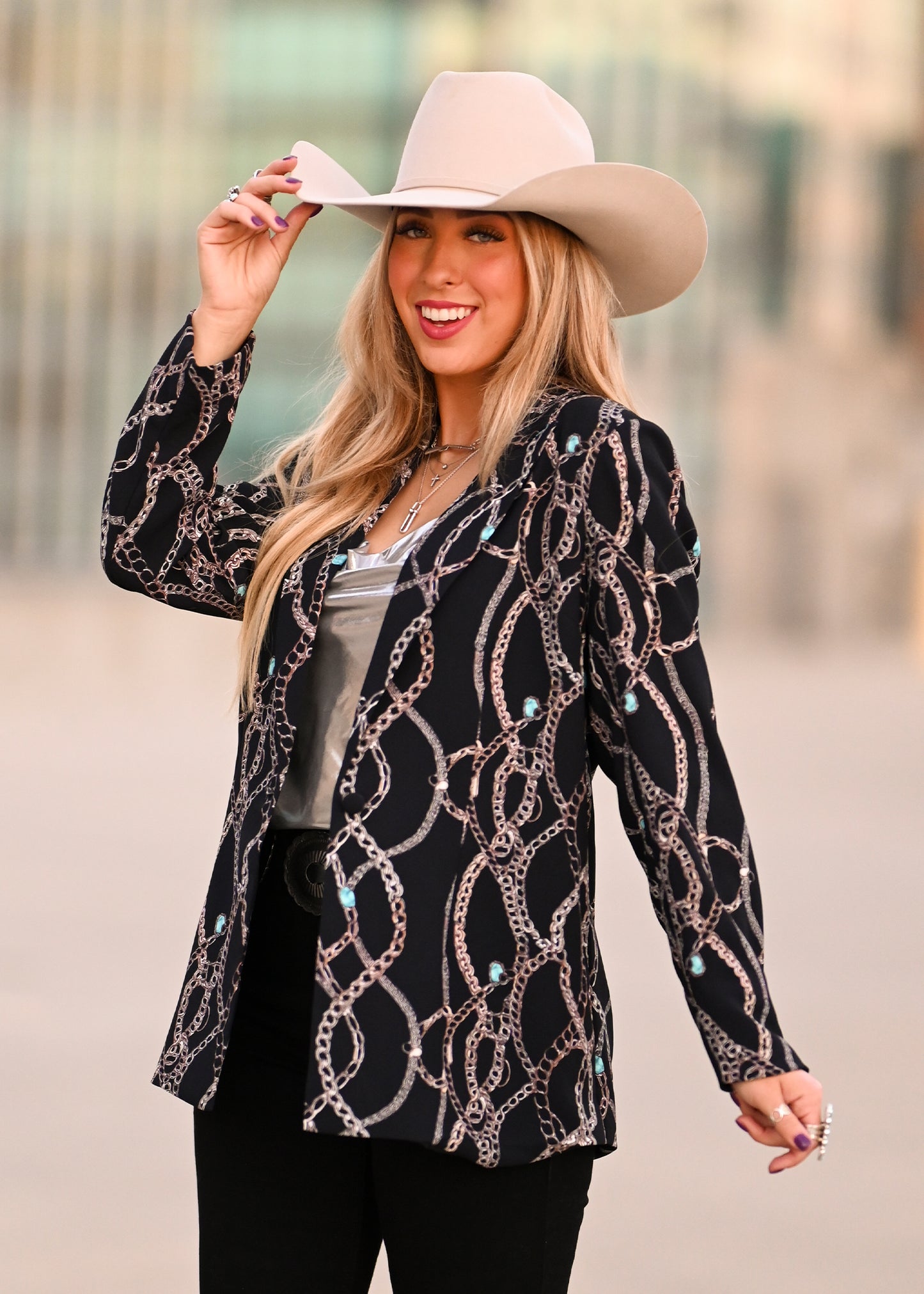 A smiling woman with long blonde hair wears a light cowboy hat, a Rock & Roll Denim Chain Print Blazer, a silver top, and black pants while standing outdoors holding the brim of her hat.