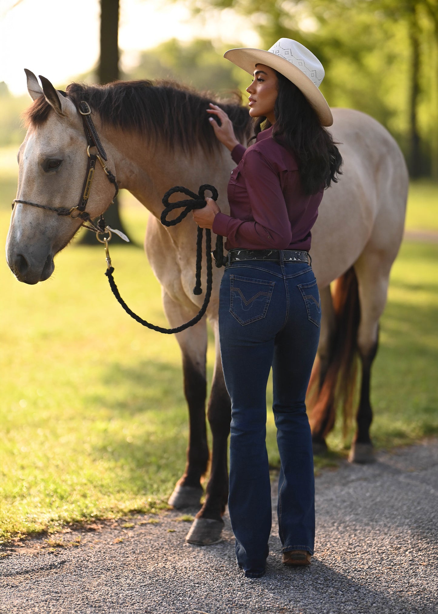 A woman in a cowboy hat, burgundy long-sleeve shirt, and blue jeans stands beside a light-colored horse on a gravel path in a sunny outdoor setting.