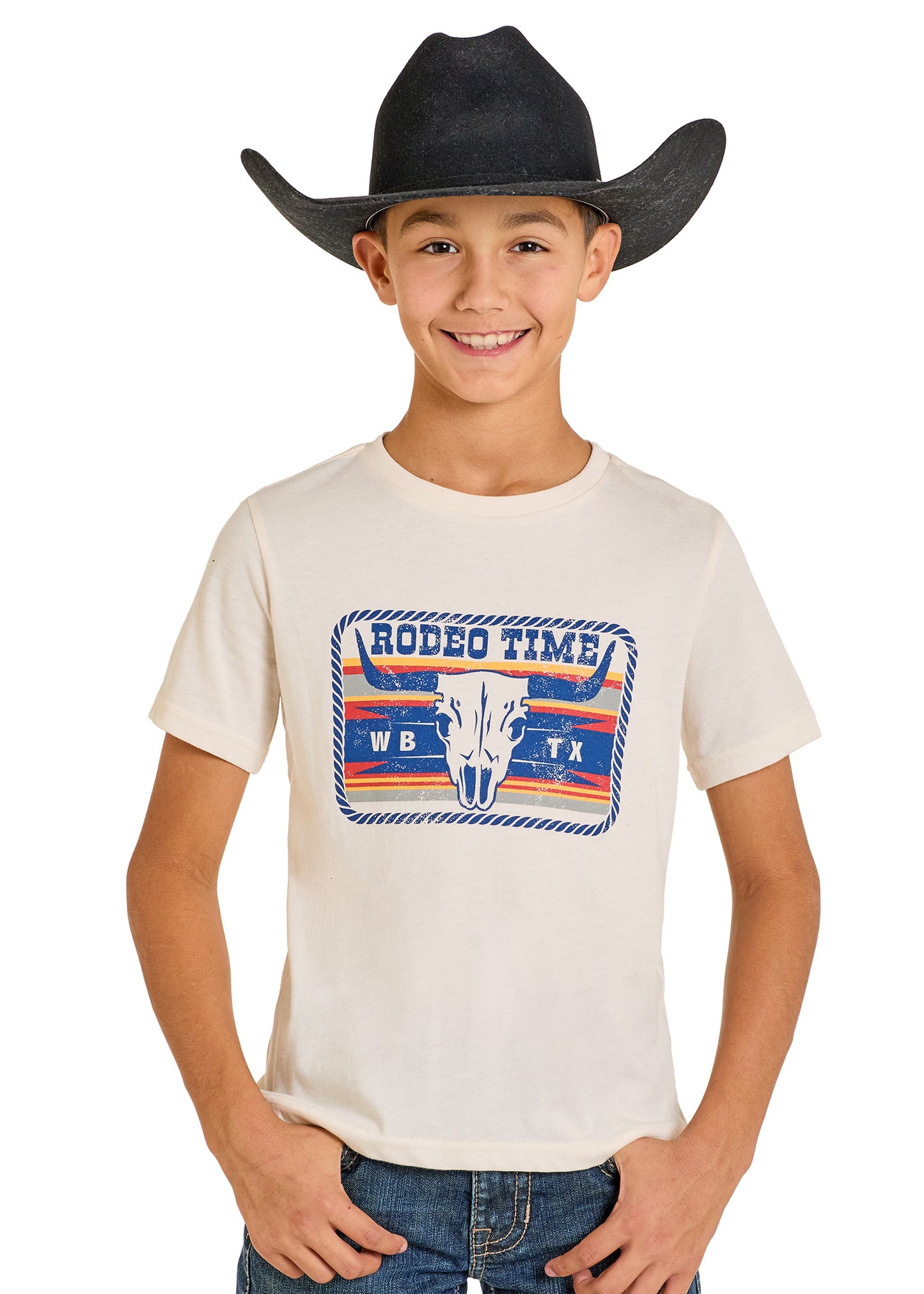 A smiling boy in a black cowboy hat, blue jeans, and a Rock & Roll Denim Boys' Dale Brisby Rodeo Time Buckle Graphic Tee featuring a bull skull stands against a plain white background.