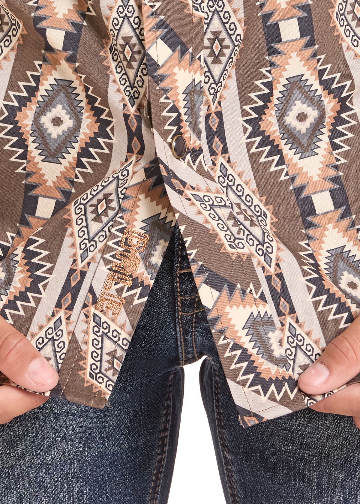 Close-up of a patterned button-up shirt being held open by hands, showing dark blue jeans underneath. The shirt features a geometric Southwestern-style print in brown, beige, black, and white.