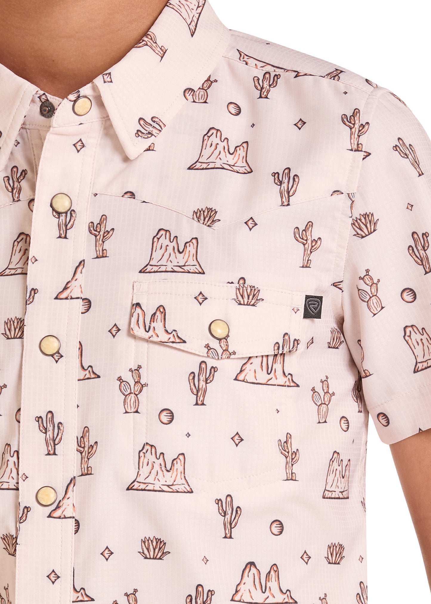 A close-up of a short-sleeve button-up shirt worn by a model, featuring a light pink base with a brown desert landscape print of mountains, cacti, and stars.
