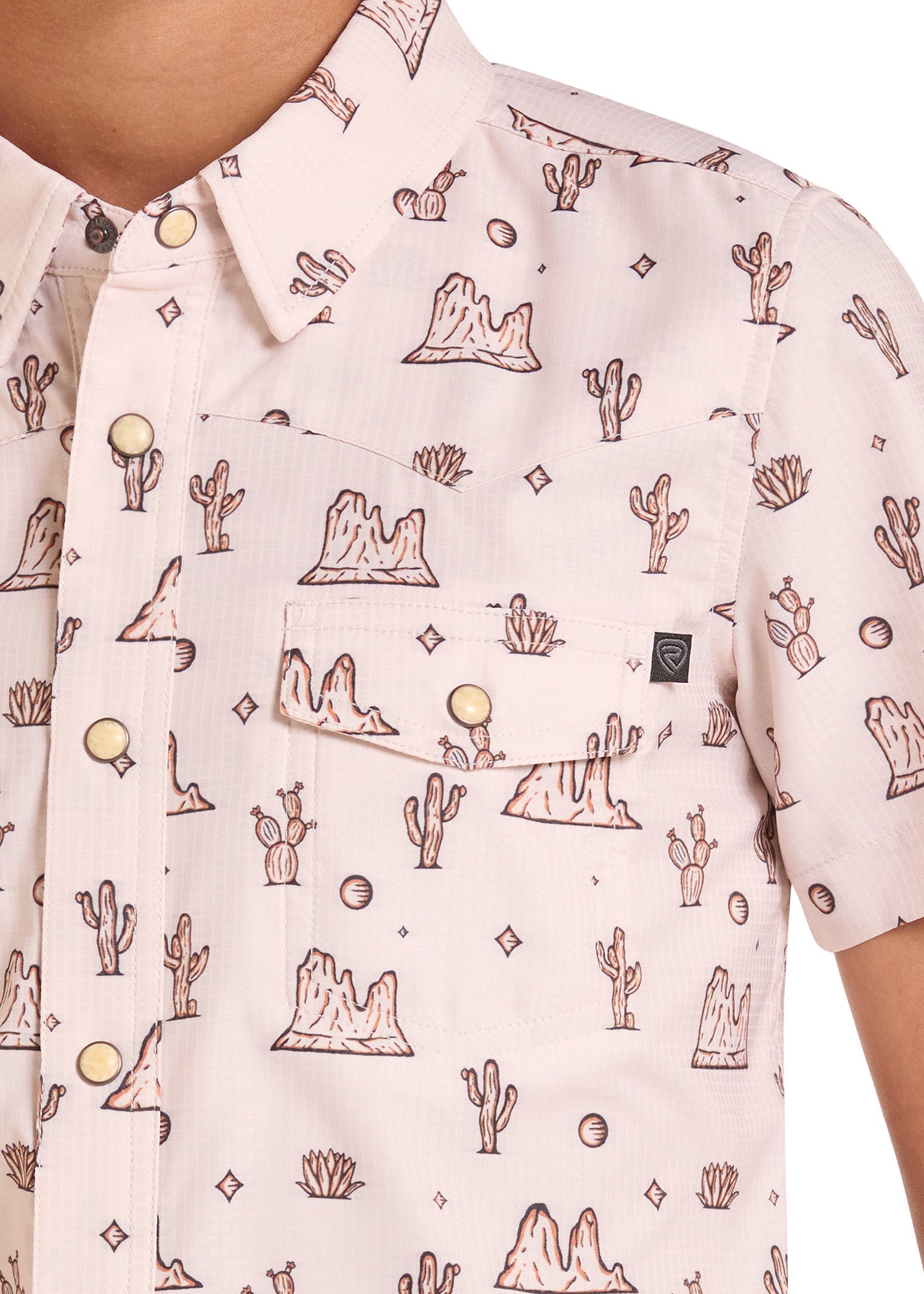 A close-up of a short-sleeve button-up shirt worn by a model, featuring a light pink base with a brown desert landscape print of mountains, cacti, and stars.